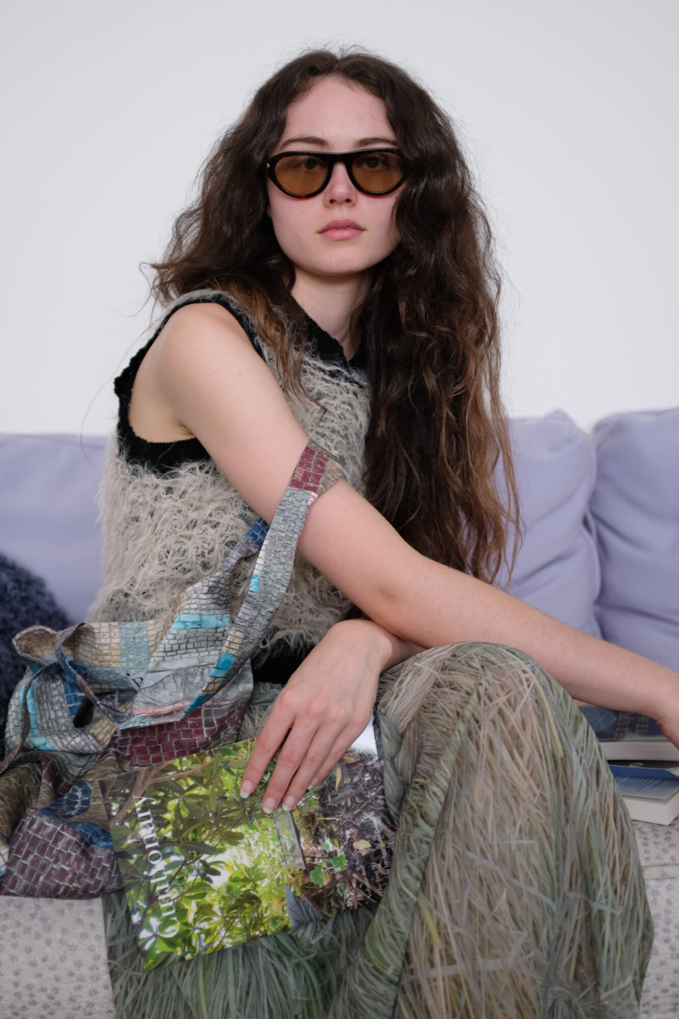Young woman with long curly hair wearing sunglasses, sitting on a sofa with a colorful bag and patterned pants.