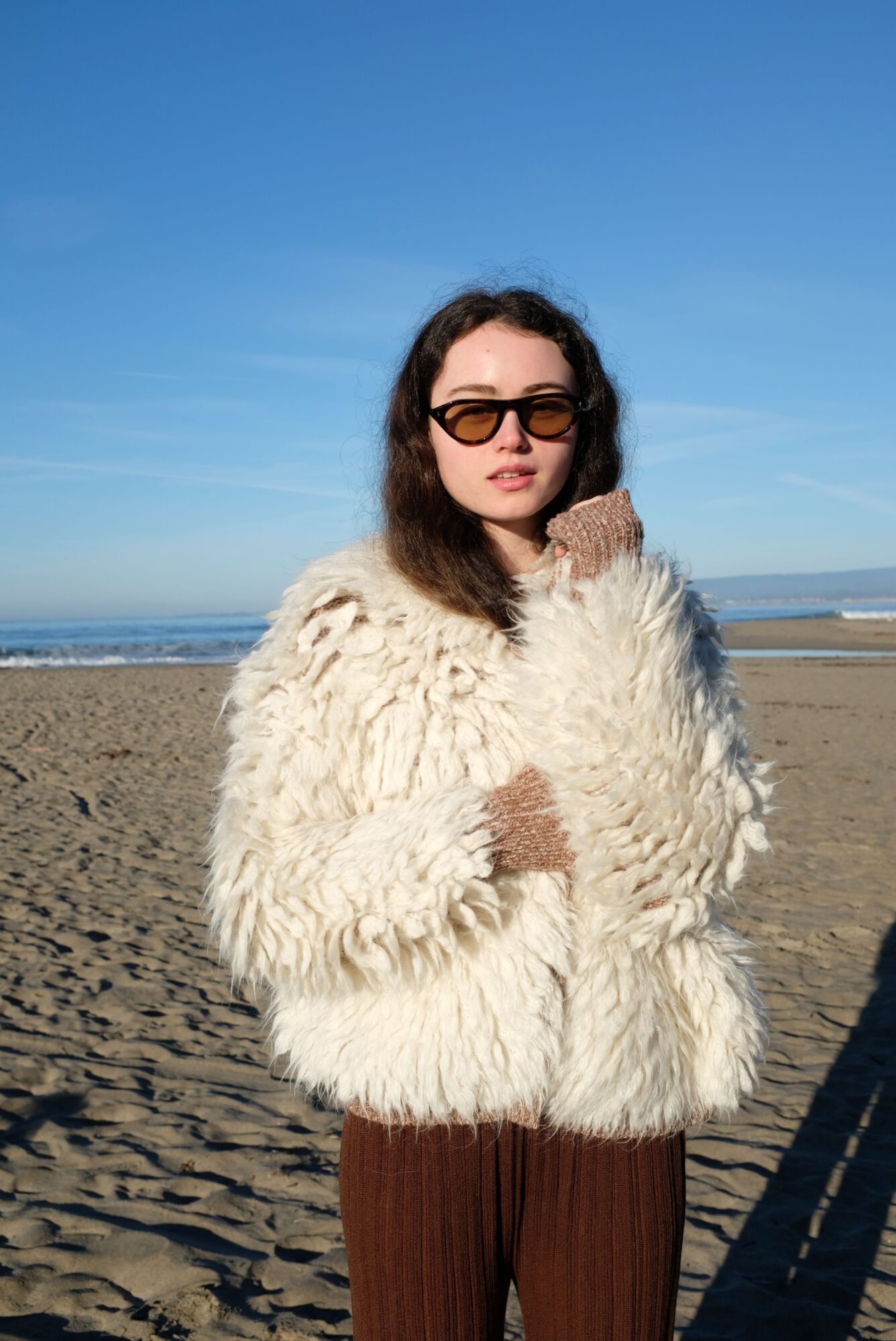 Woman in sunglasses and a fluffy coat standing on a sandy beach with blue sky and ocean in background.