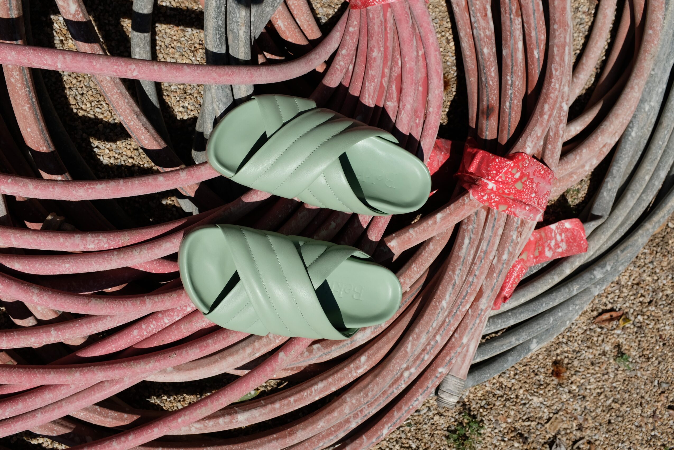 Pair of green slip-on shoes on coiled pink and gray hoses on ground, with dirt nearby.