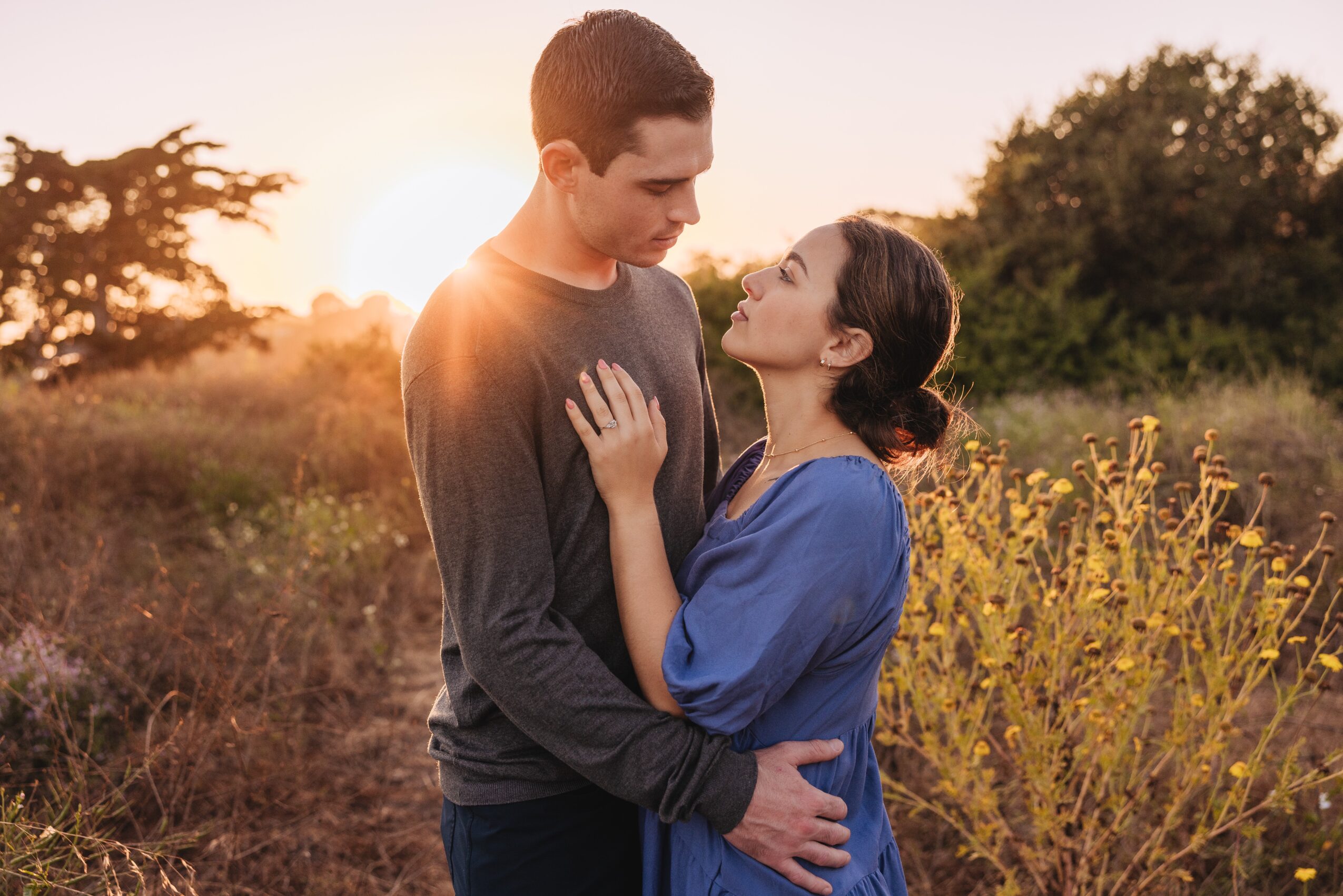 A man and woman standing close outdoors during sunset, gazing at each other, with trees and yellow flowers in the background.