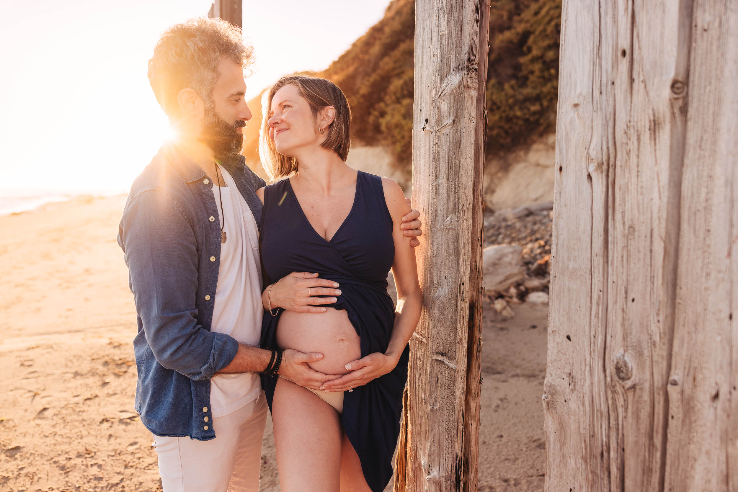 A couple stands outdoors near a wooden structure, with the sun setting behind them, embracing and smiling at each other.