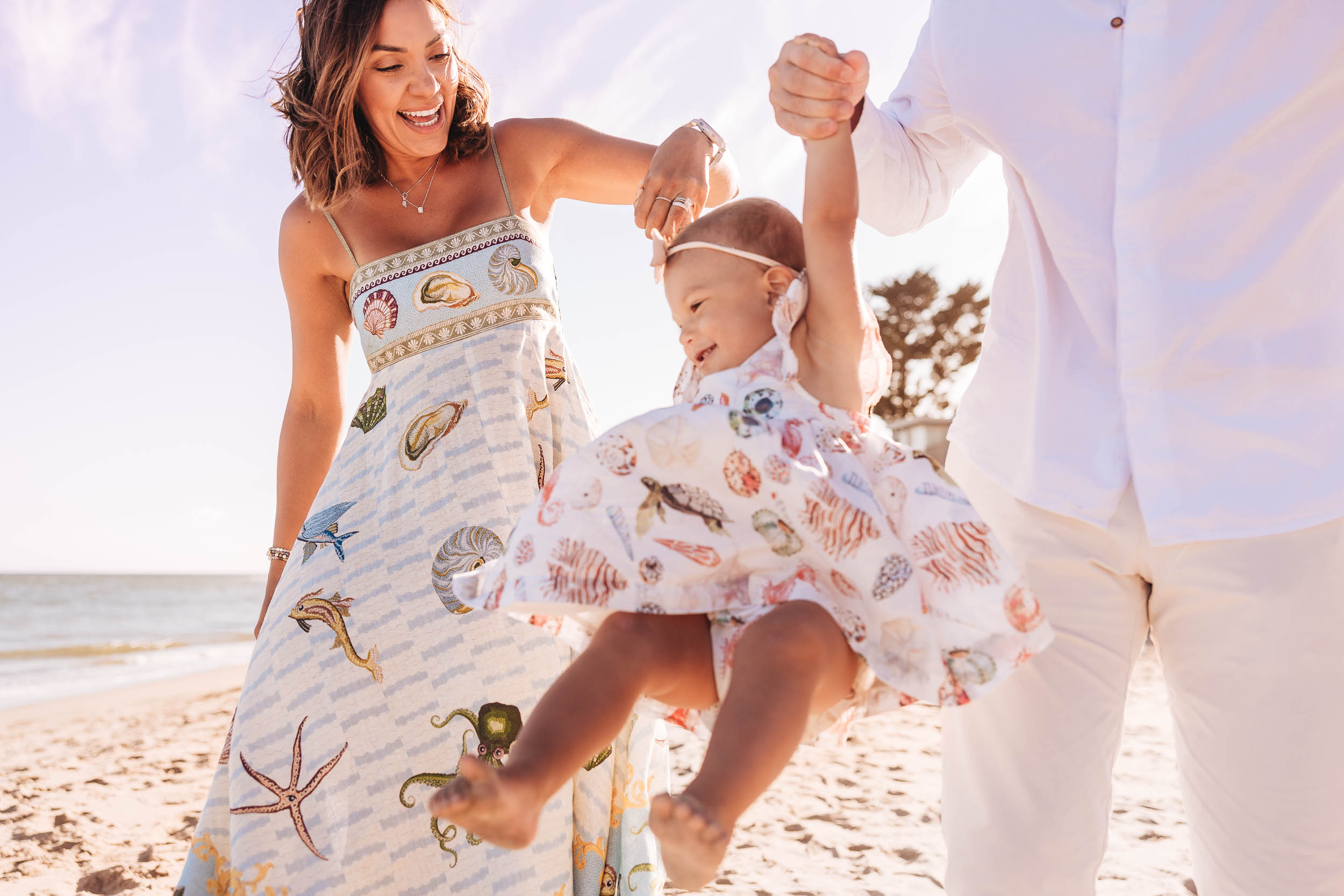 Woman and man holding a child in a dress on a beach, smiling and playing, with ocean and sky in background.
