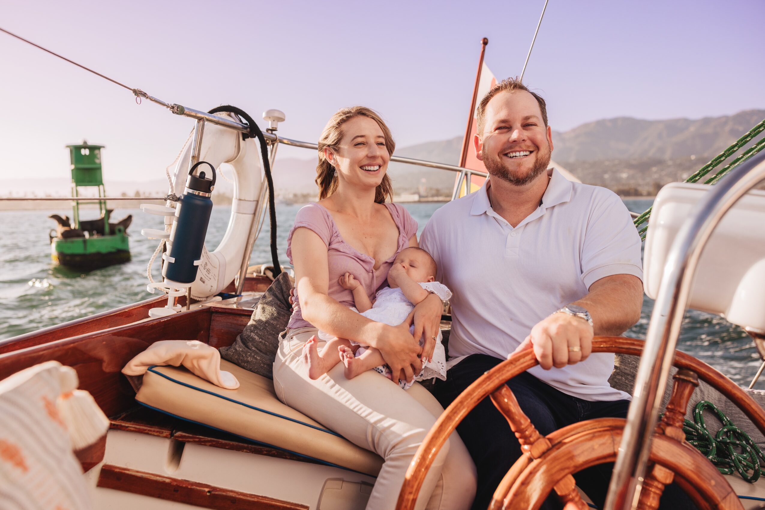 A smiling family of three on a boat, with water and mountains in the background, enjoying a sunny day.