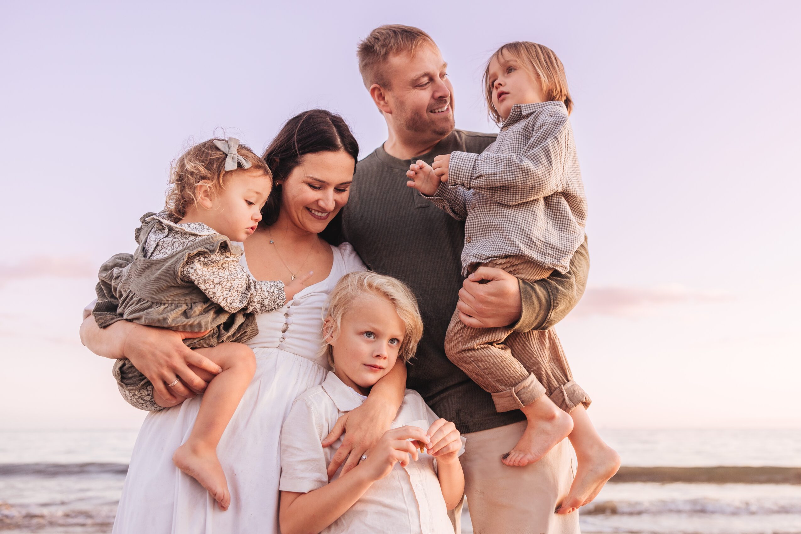 Family of five outdoors at the beach, smiling and hugging, with a man, woman, and three children, during sunset.