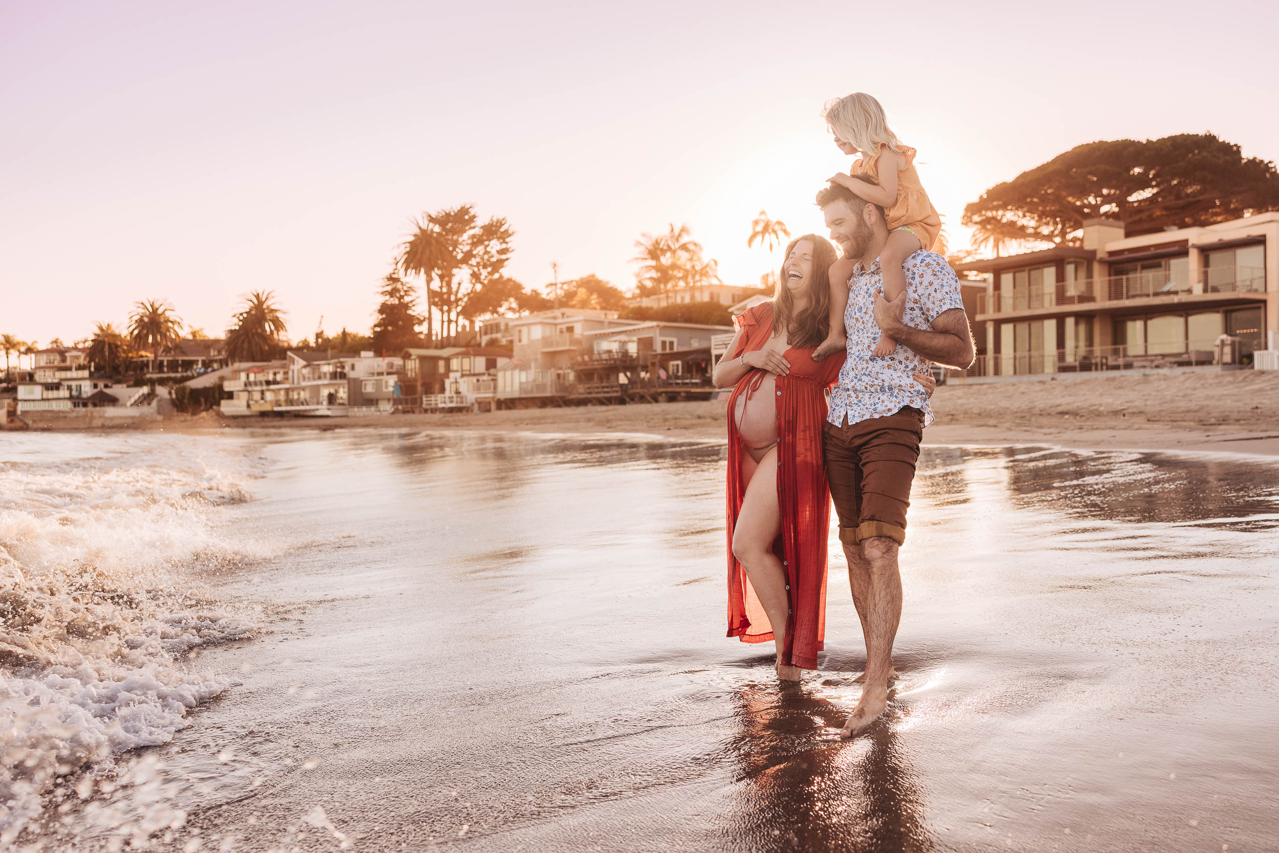 Family walking along the beach at sunset, with houses and trees in the background, and a child on a parent's shoulders.