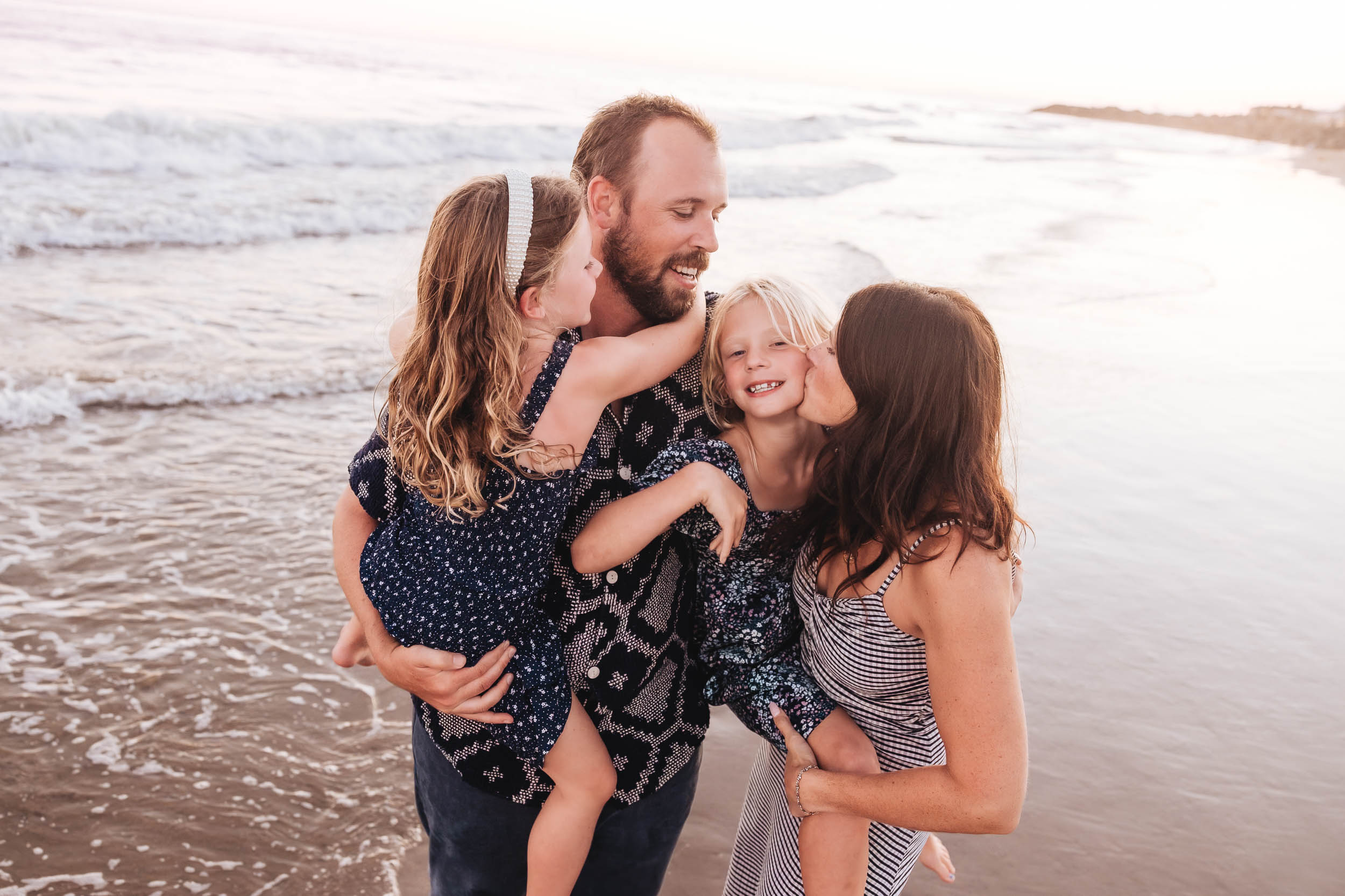 A family of four, including a man, woman, and two young girls, stands on a beach with waves in the background.