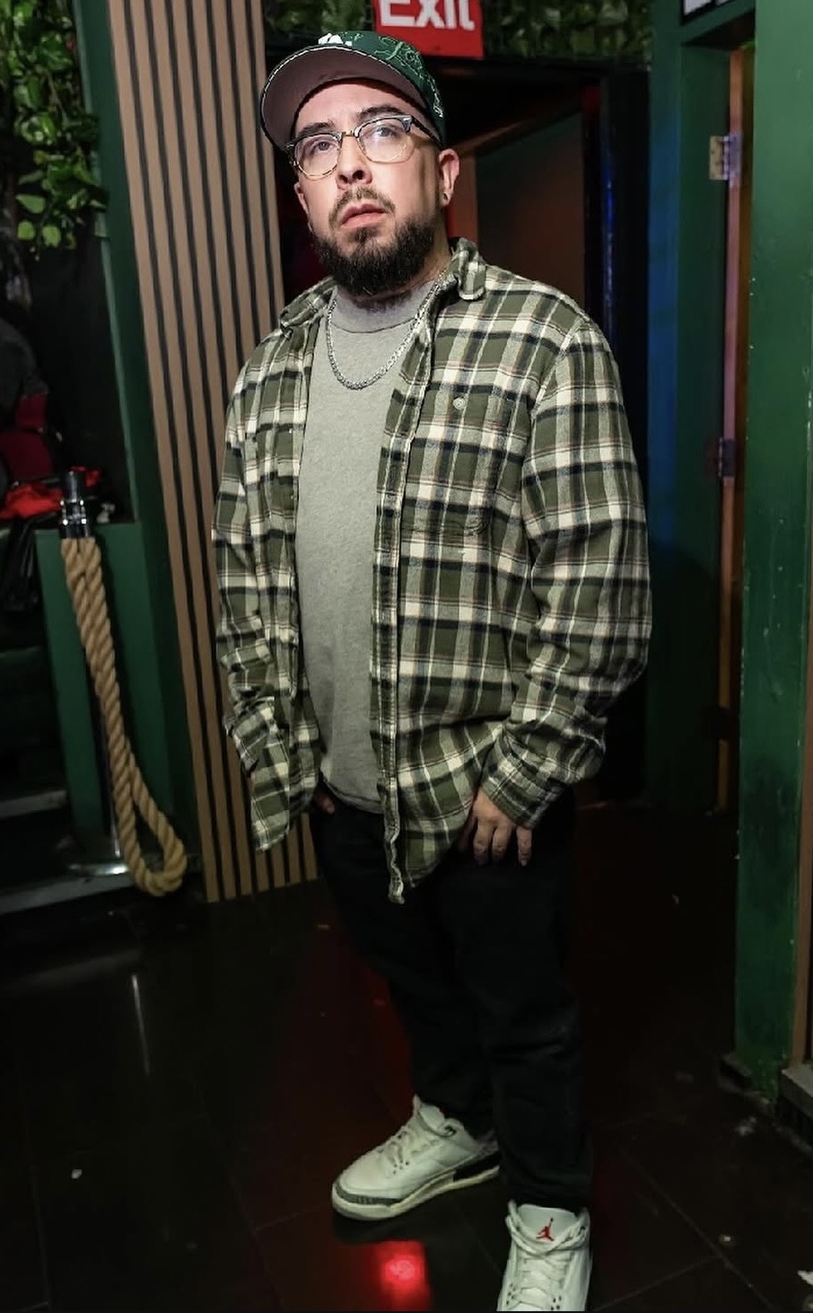 Young man with glasses and beard wearing a plaid shirt, gray t-shirt, and white sneakers, standing indoors.