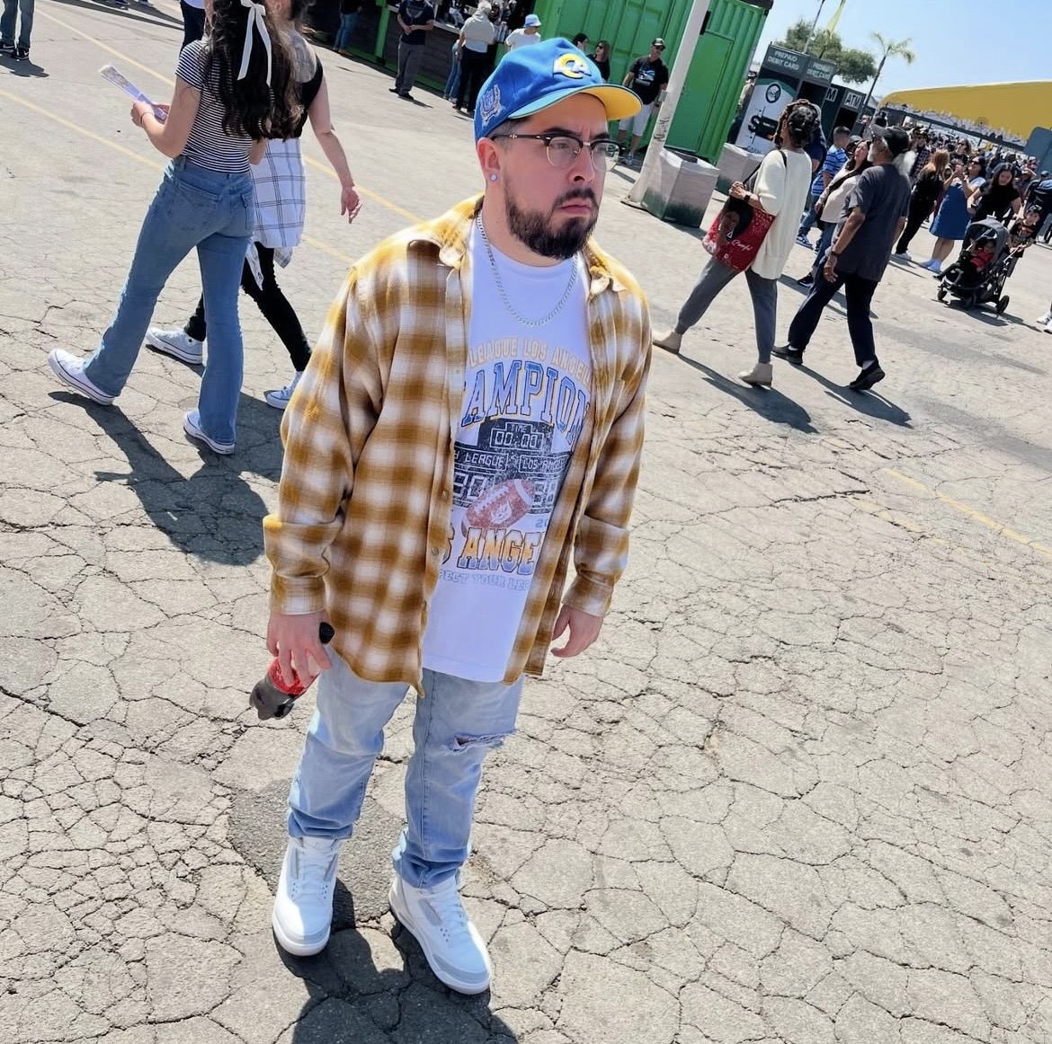 Man with glasses, beard, blue cap, yellow plaid shirt, white t-shirt, light jeans, white sneakers, standing on cracked pavement with people in background.