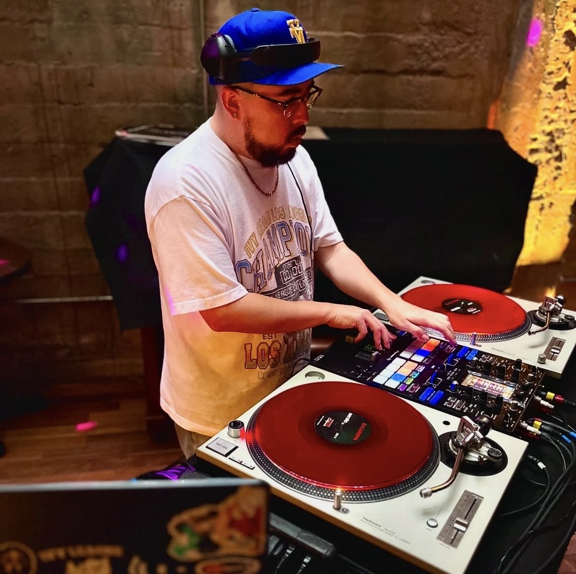 DJ wearing a blue cap and glasses, operating turntables and mixer in a dimly lit venue.