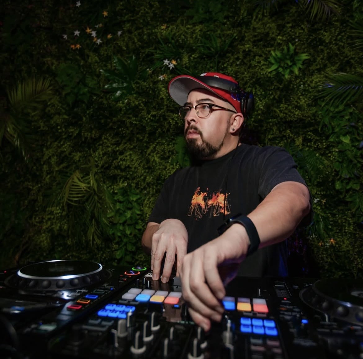 DJ wearing glasses and a cap, operating a DJ controller with a green leafy background.