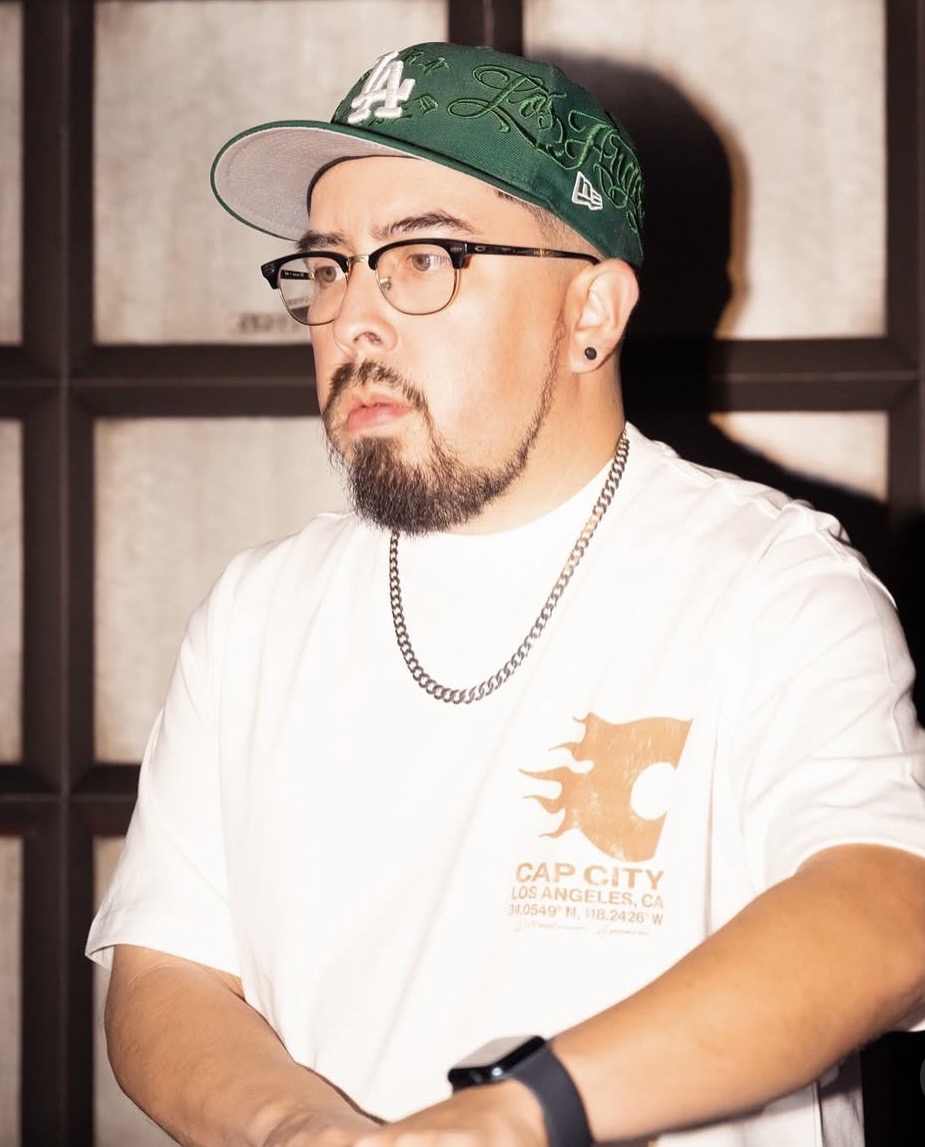 Man with glasses, beard, and earrings wearing a white T-shirt, green cap, and chain, sitting indoors.