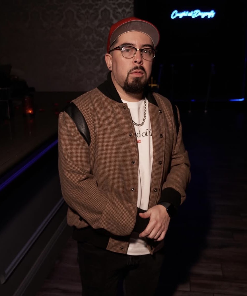 Man with glasses, beard, red cap, brown jacket, standing indoors with dark background and neon sign, looking at camera.