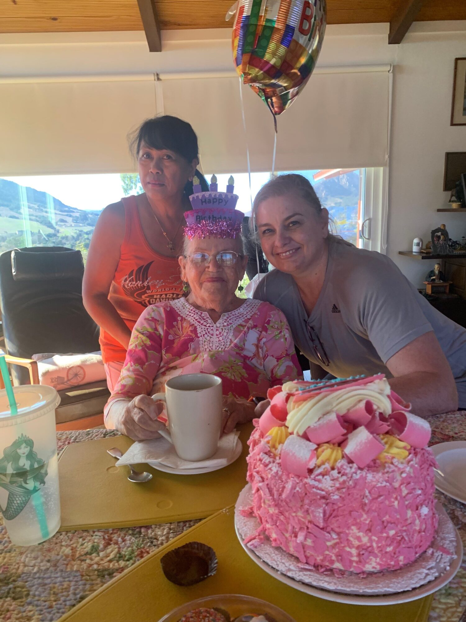 Two women celebrate a birthday with a pink cake topped with strawberries and a person holding a colorful balloon.