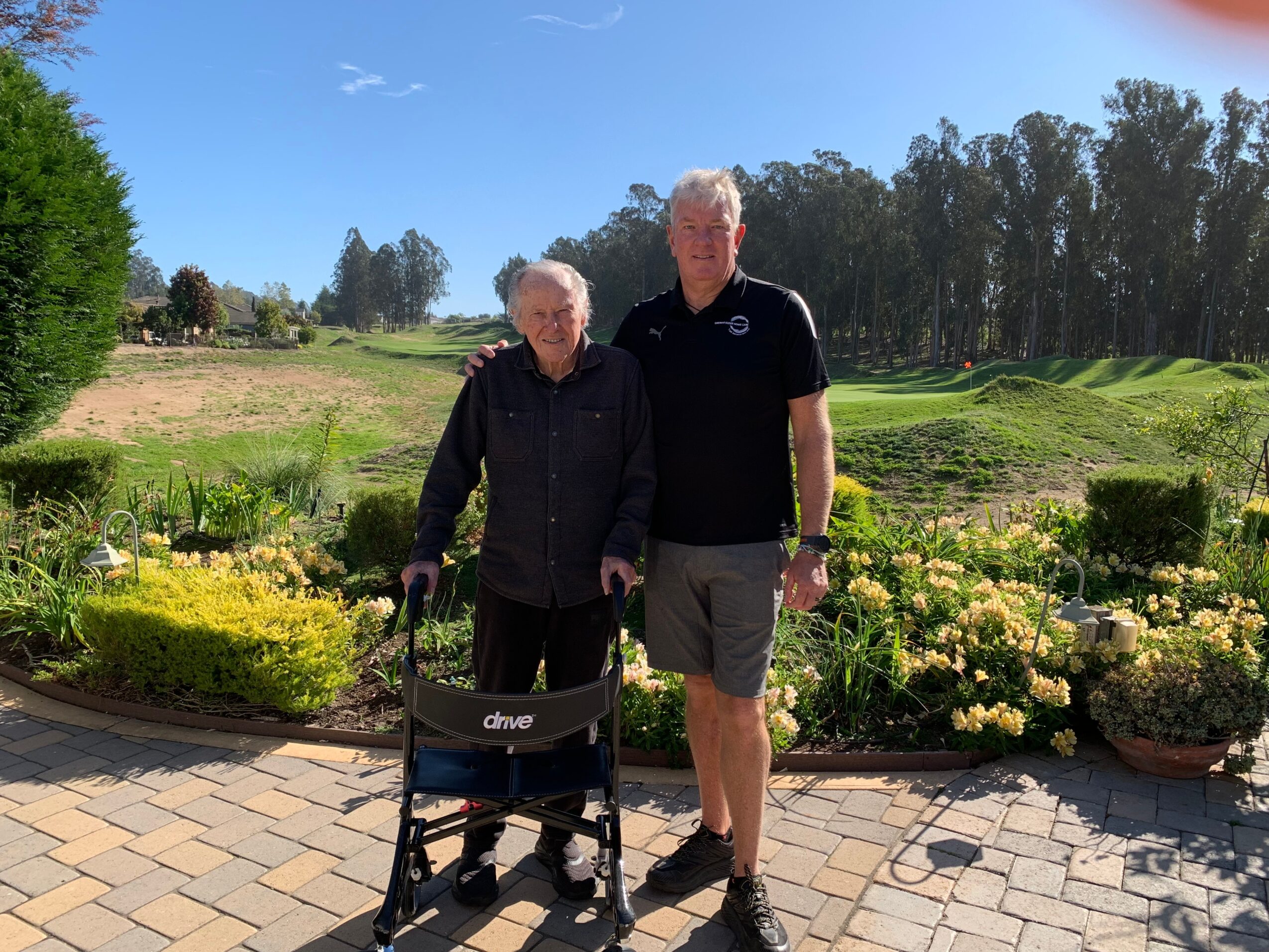 Two people standing outdoors on a paved area with a garden and trees in the background, one using a walker.