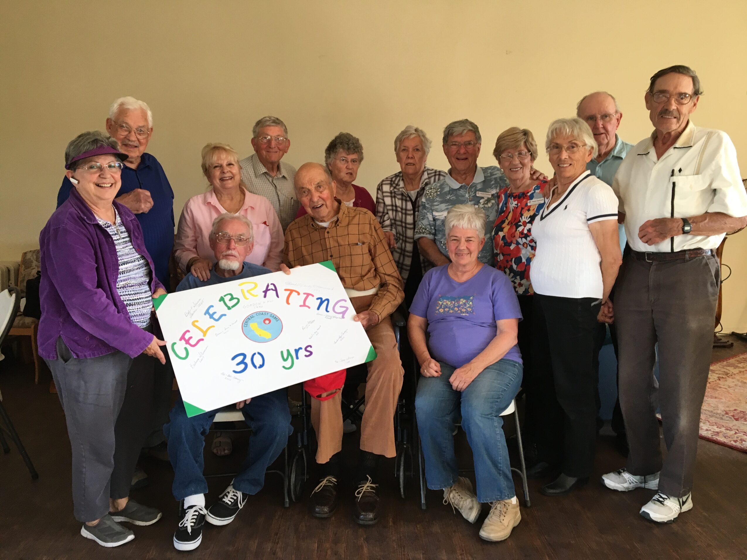 Group of elderly people holding a colorful sign celebrating 30 years, standing indoors against a plain wall.