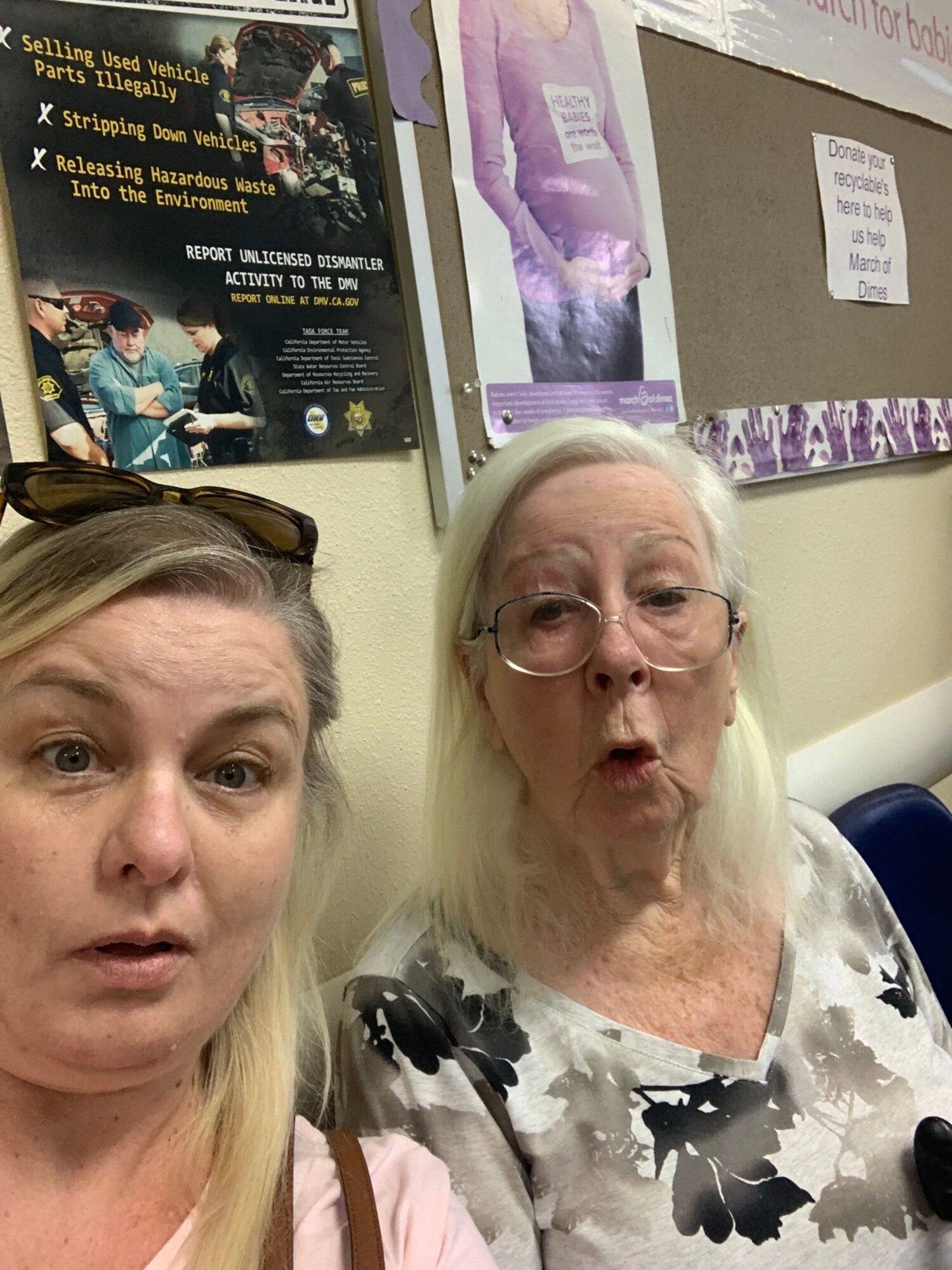 Two women with light hair and glasses in a room with posters and a purple object on the wall.