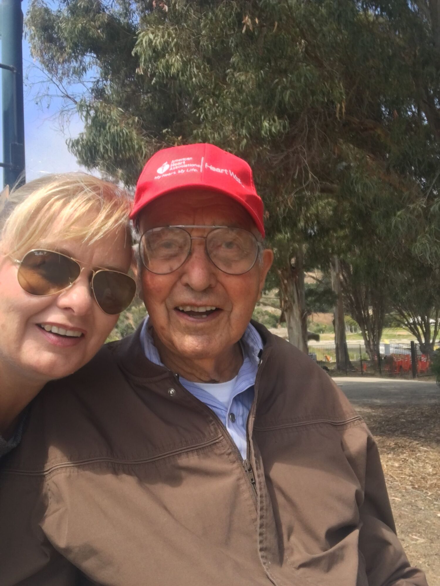 Two people outdoors, one wearing a red cap and glasses, the other with sunglasses, smiling, trees in background.