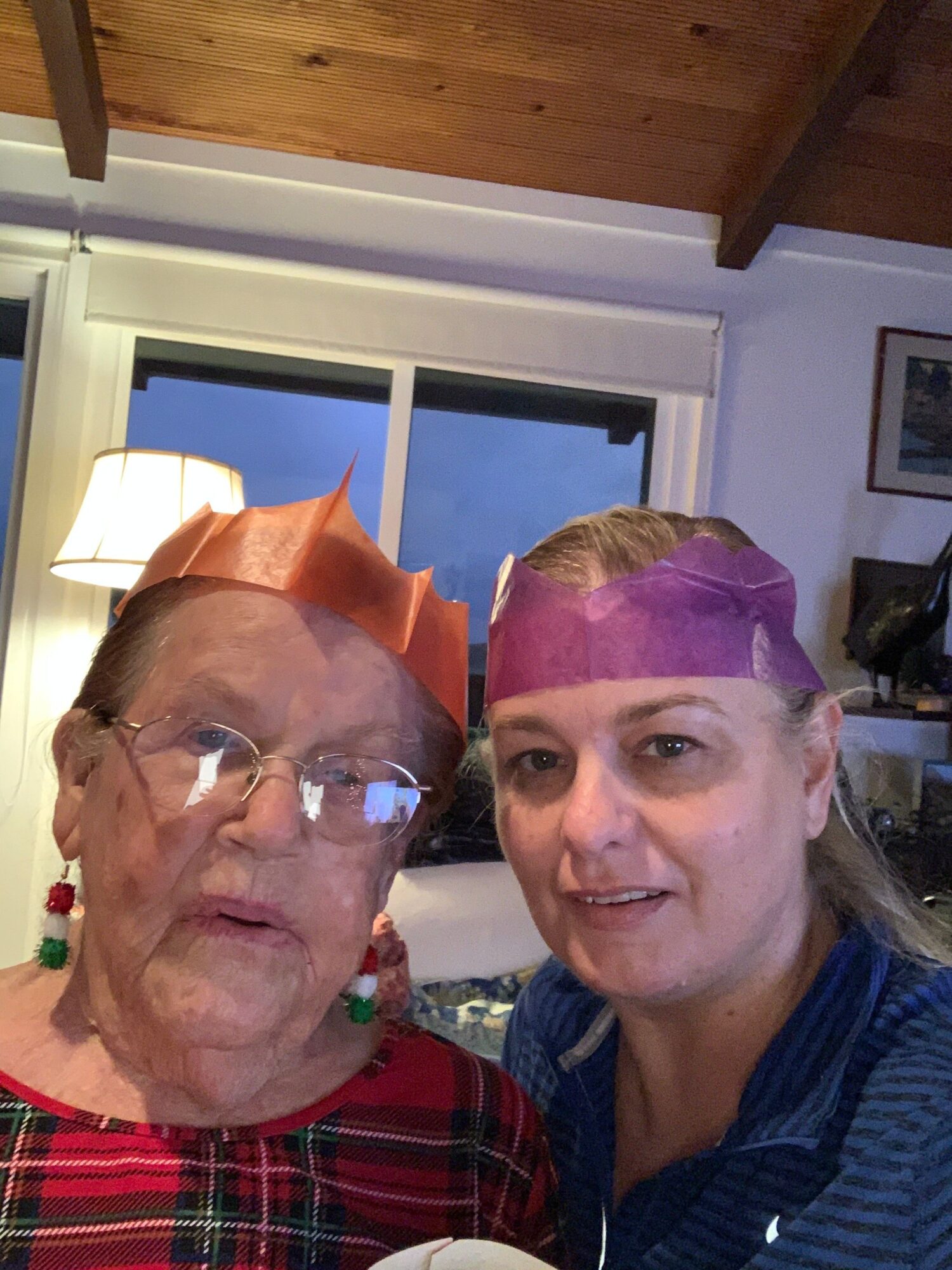 Two women smiling indoors near a window, one wearing glasses and a red headscarf, the other with a purple headscarf.