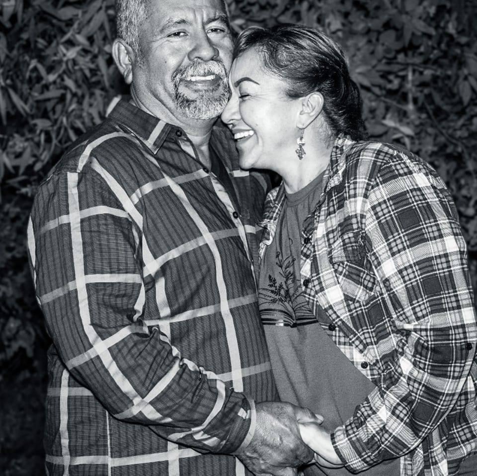 Two people smiling and holding hands, standing close together outdoors, wearing plaid shirts.
