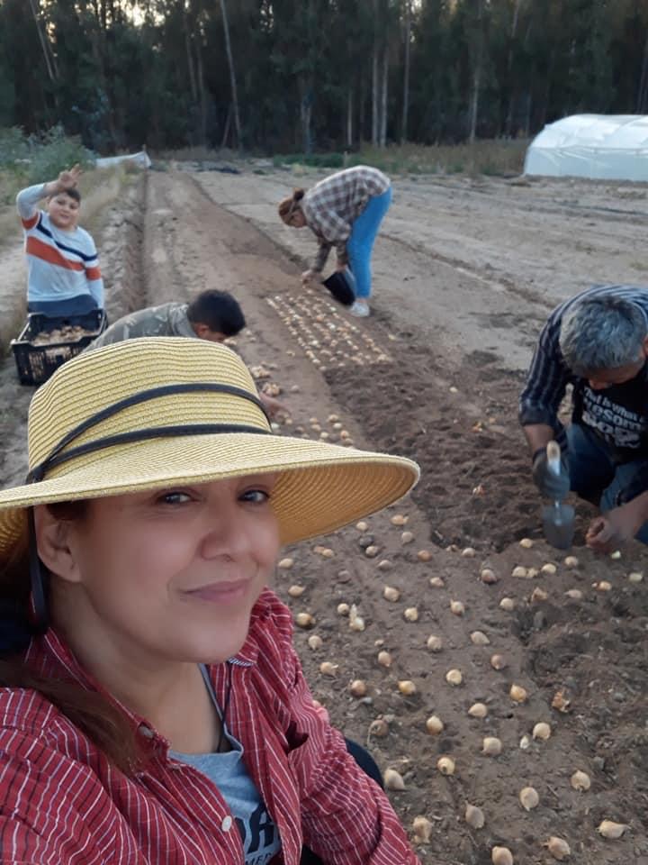 Woman wearing a wide-brimmed hat taking a selfie in a farm field with children planting seeds in the background.