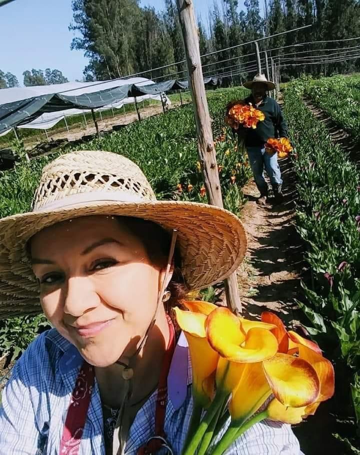 Woman wearing a straw hat smiling in a garden with a person in the background holding flowers.