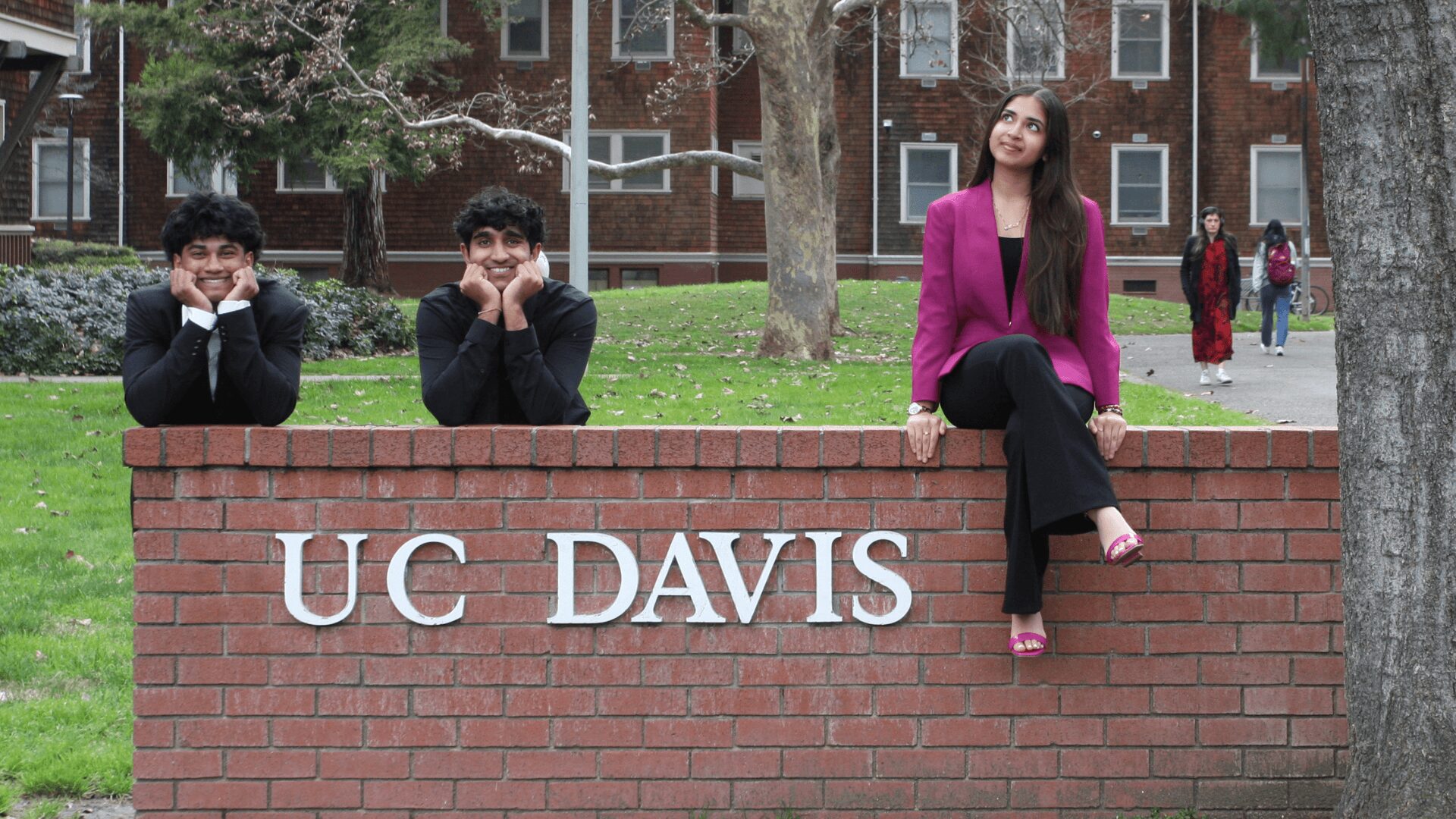 Three people sitting on a brick wall with 'UC DAVIS' sign, trees, and a building in the background.