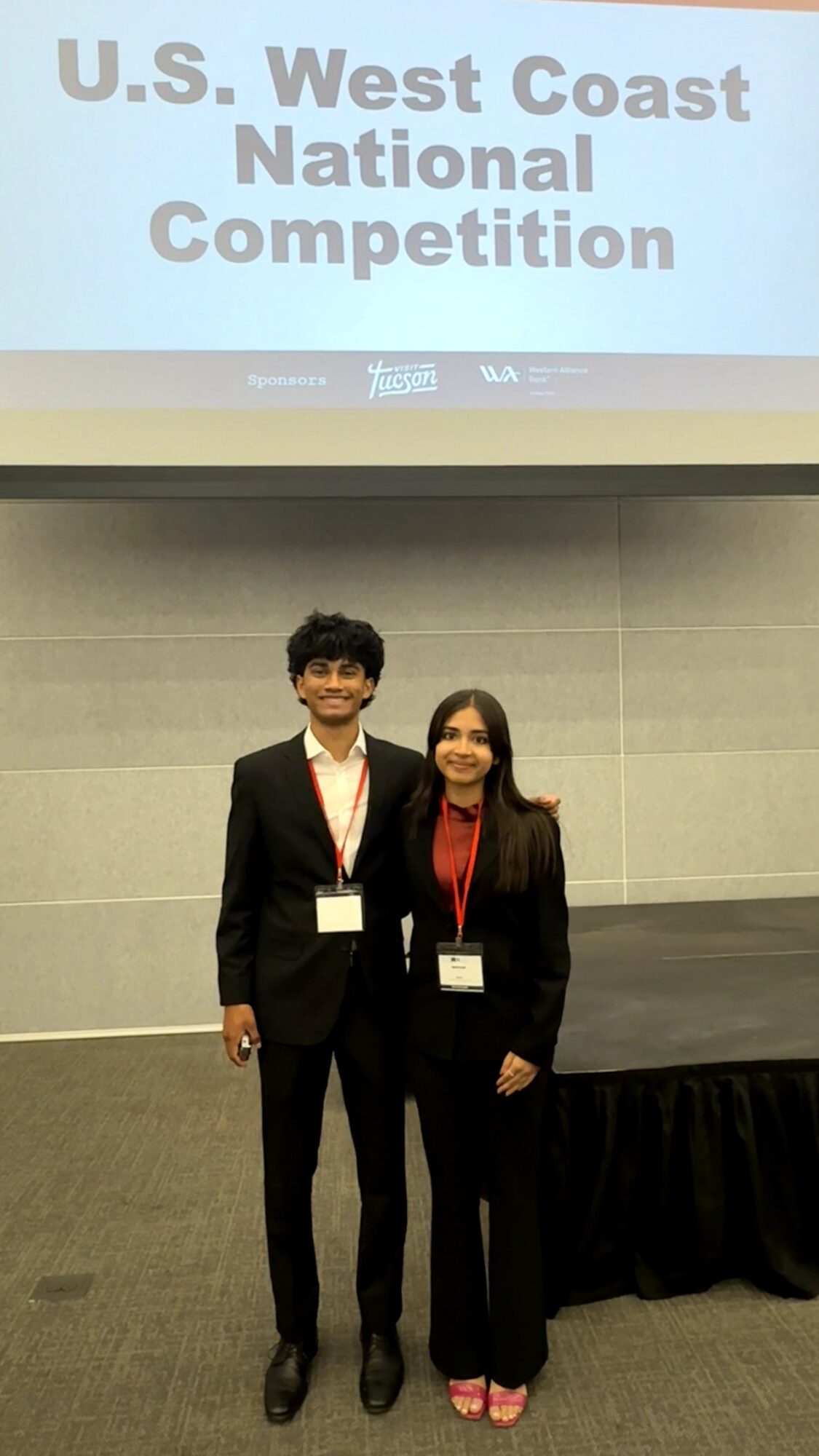 Two people in formal attire standing in front of a presentation screen that reads 'U.S. West Coast National Competition.'