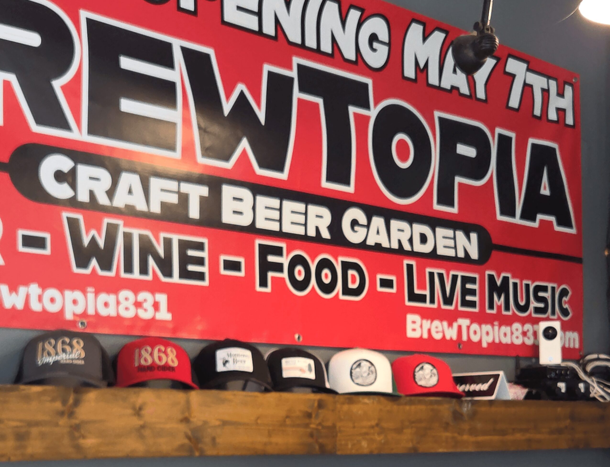 Red and black banner advertising Brewtopia craft beer garden, wine, food, and live music, with hats displayed below.