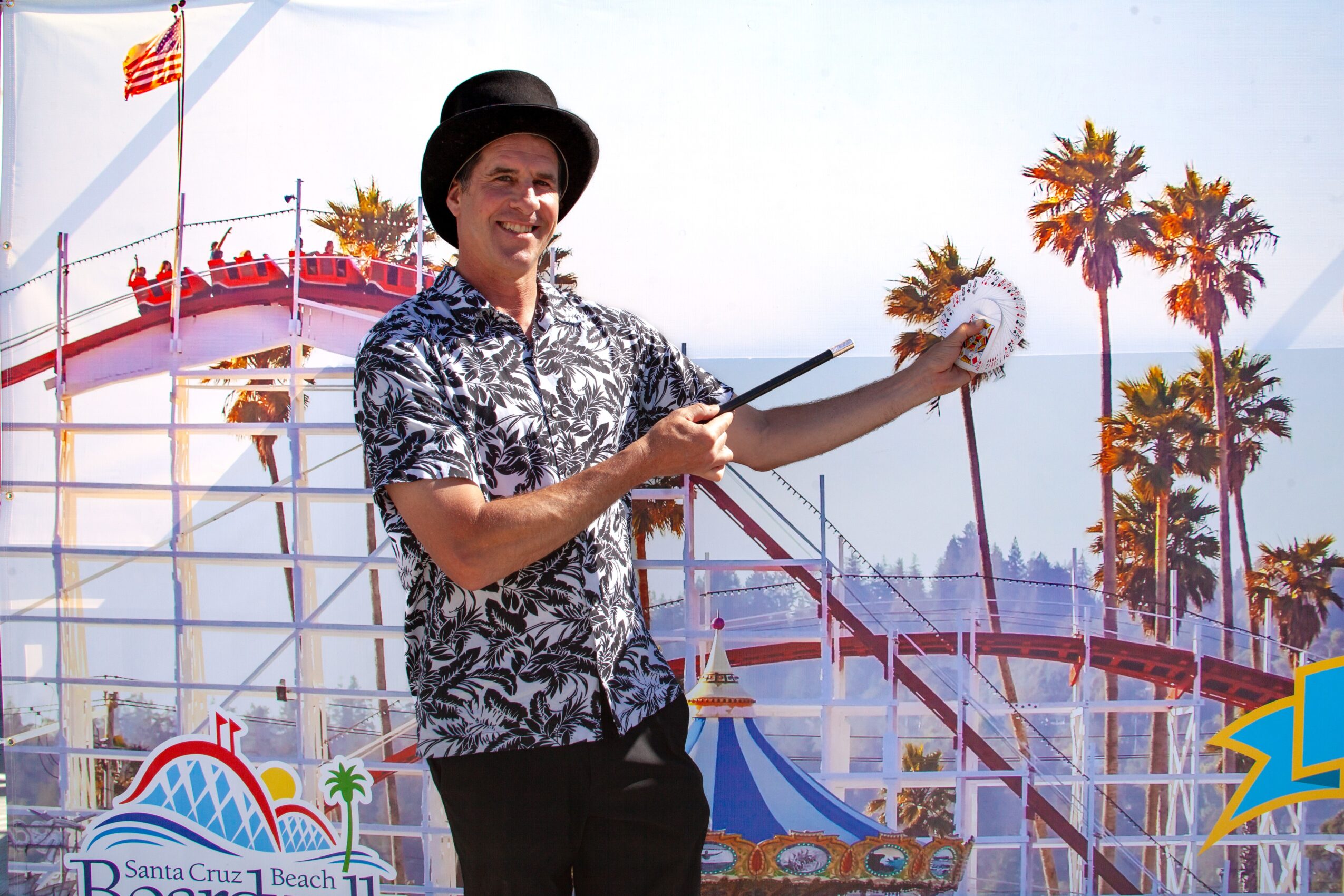 Person wearing a hat and patterned shirt holding a feather duster at a beachside amusement park with palm trees and a roller coaster in the background.