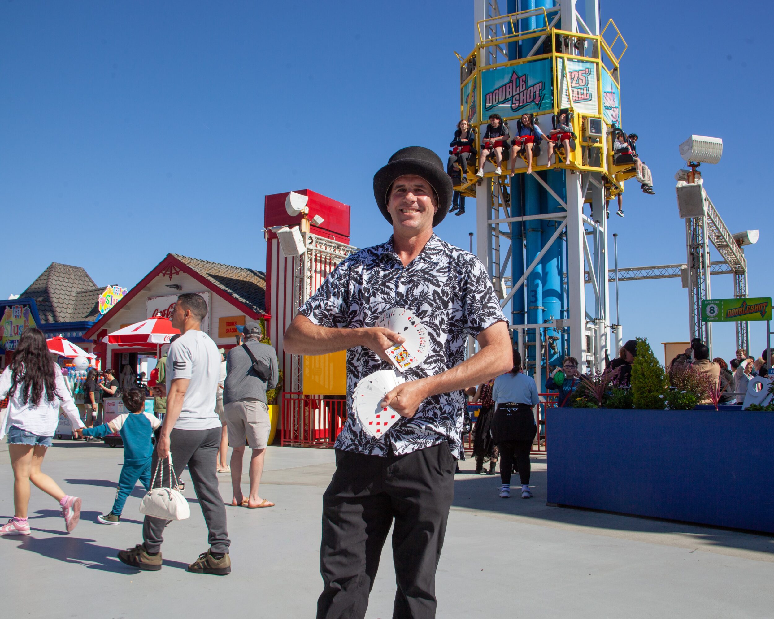 Person smiling in front of amusement park rides and visitors on a sunny day.