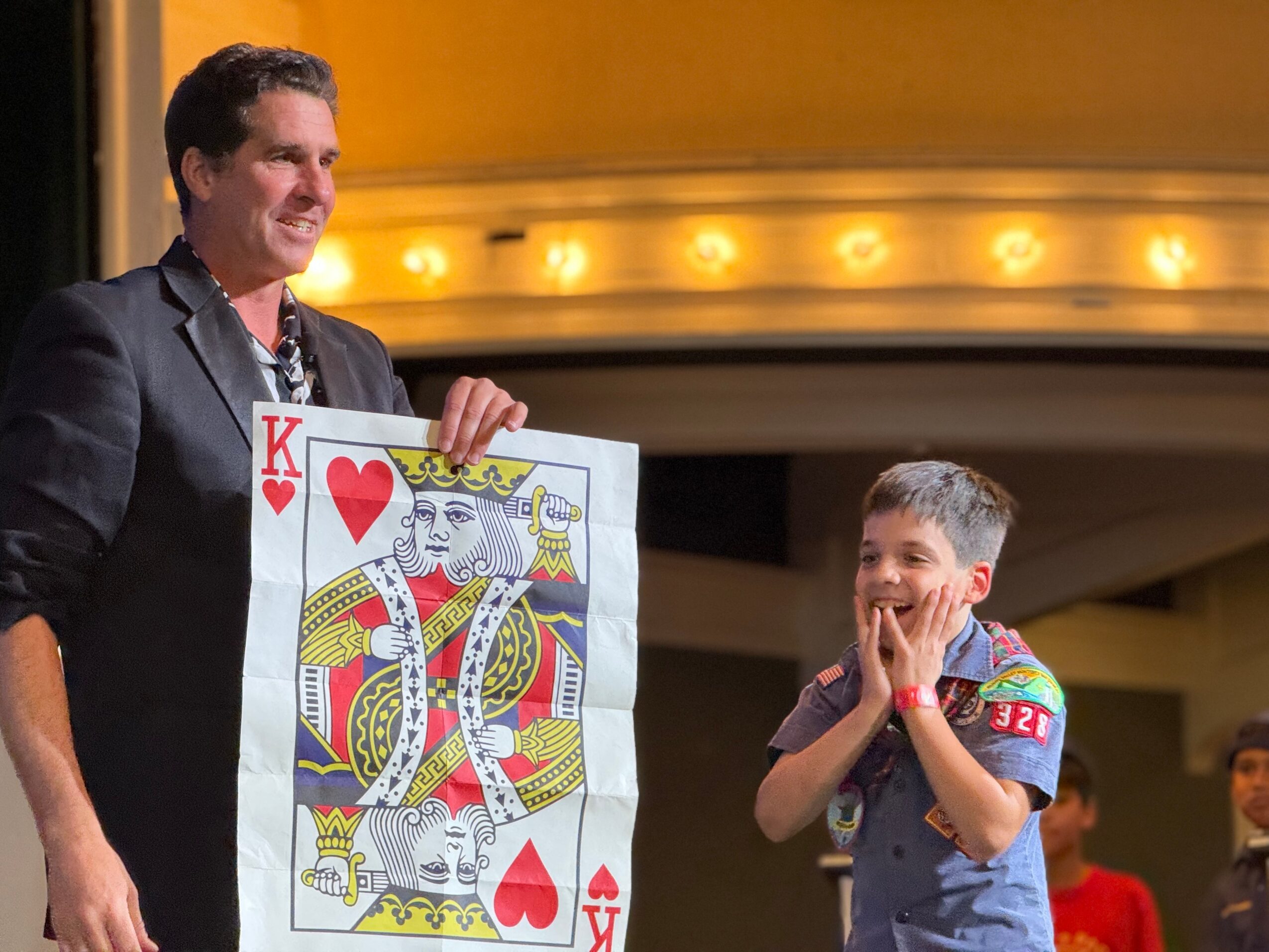Man holding a large playing card on stage, young boy reacting excitedly, theater or stage setting in background.