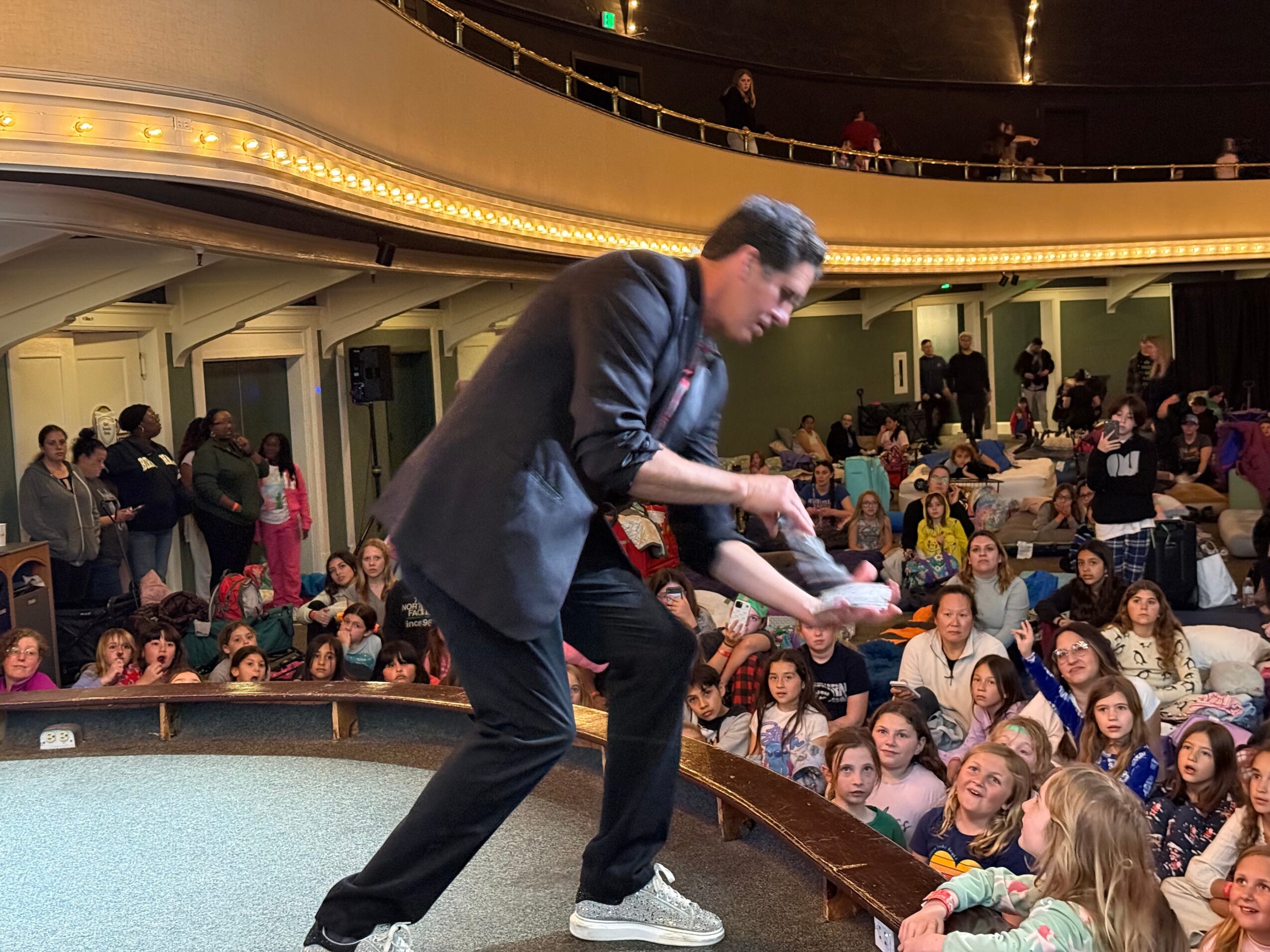 Performer in a suit interacts with children in an auditorium filled with seated audience members.