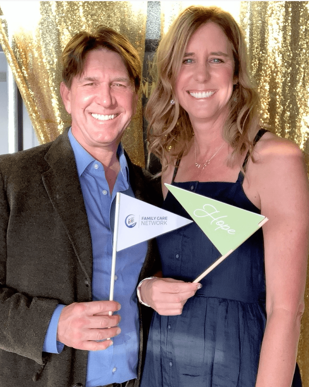 Two smiling people holding small flags, standing in front of a gold curtain backdrop.