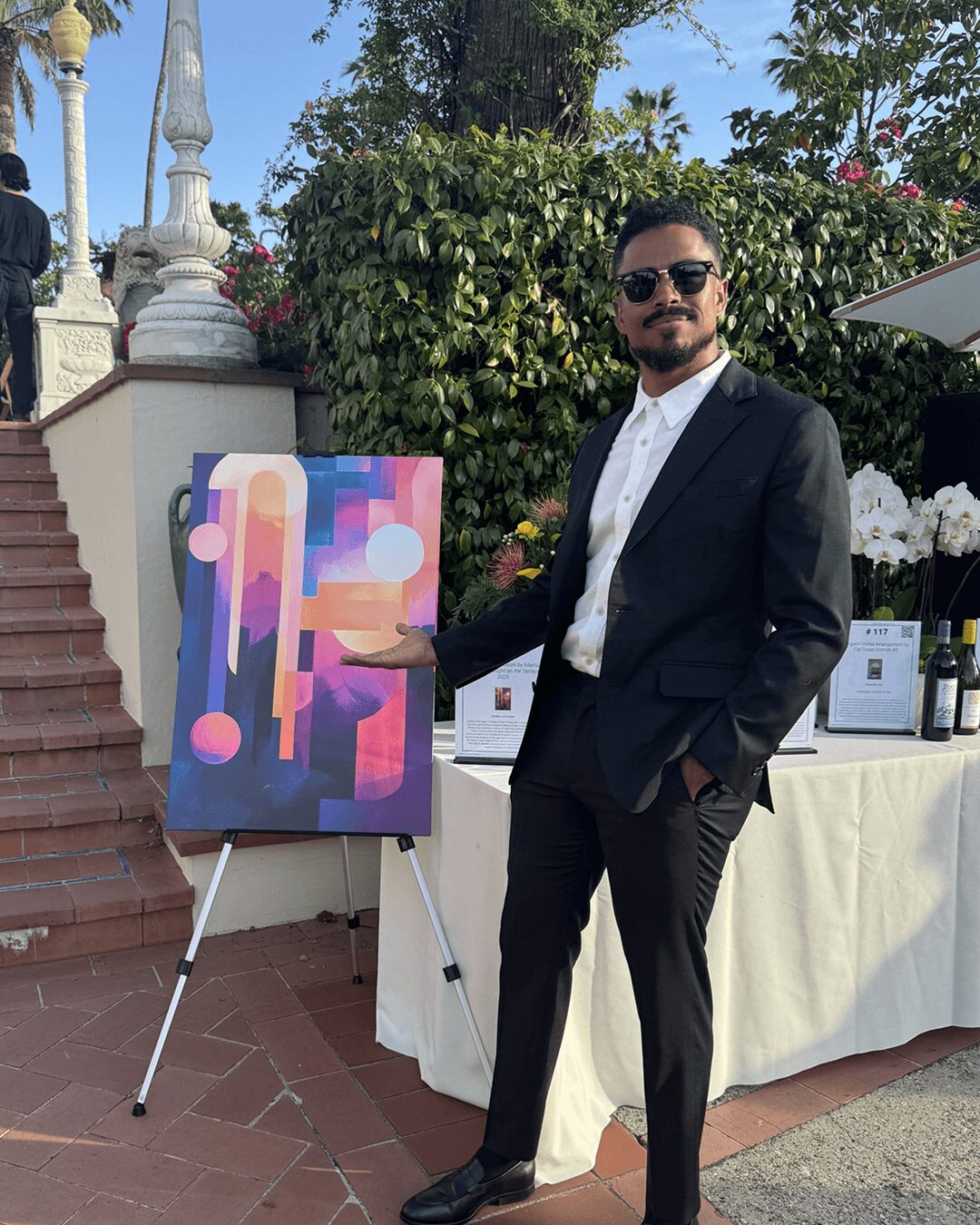 Man in a black suit and sunglasses standing outdoors next to an abstract colorful painting on an easel.