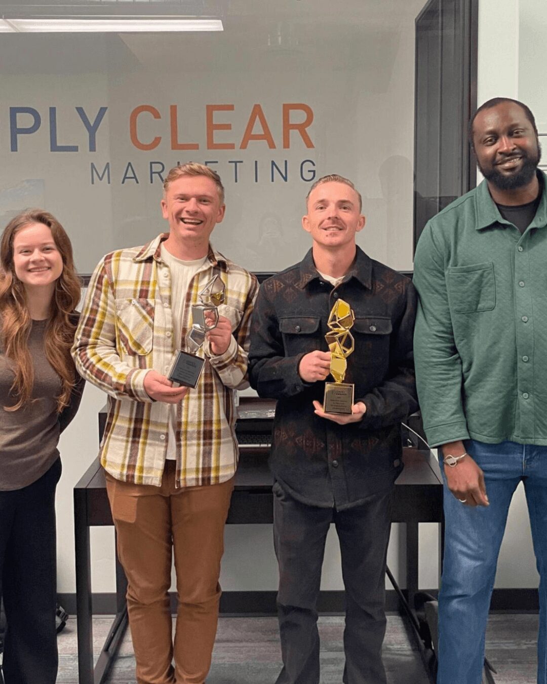Four people standing in front of a glass wall with 'CLEAR MARKETING' text, holding awards, smiling.