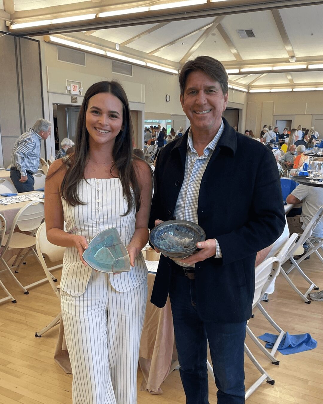 A young woman and an older man stand indoors, each holding a glass art piece, smiling at the camera. Background shows a busy event space.