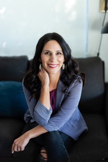 Woman with dark hair smiling, sitting on a dark sofa, hand on face, in a bright room.