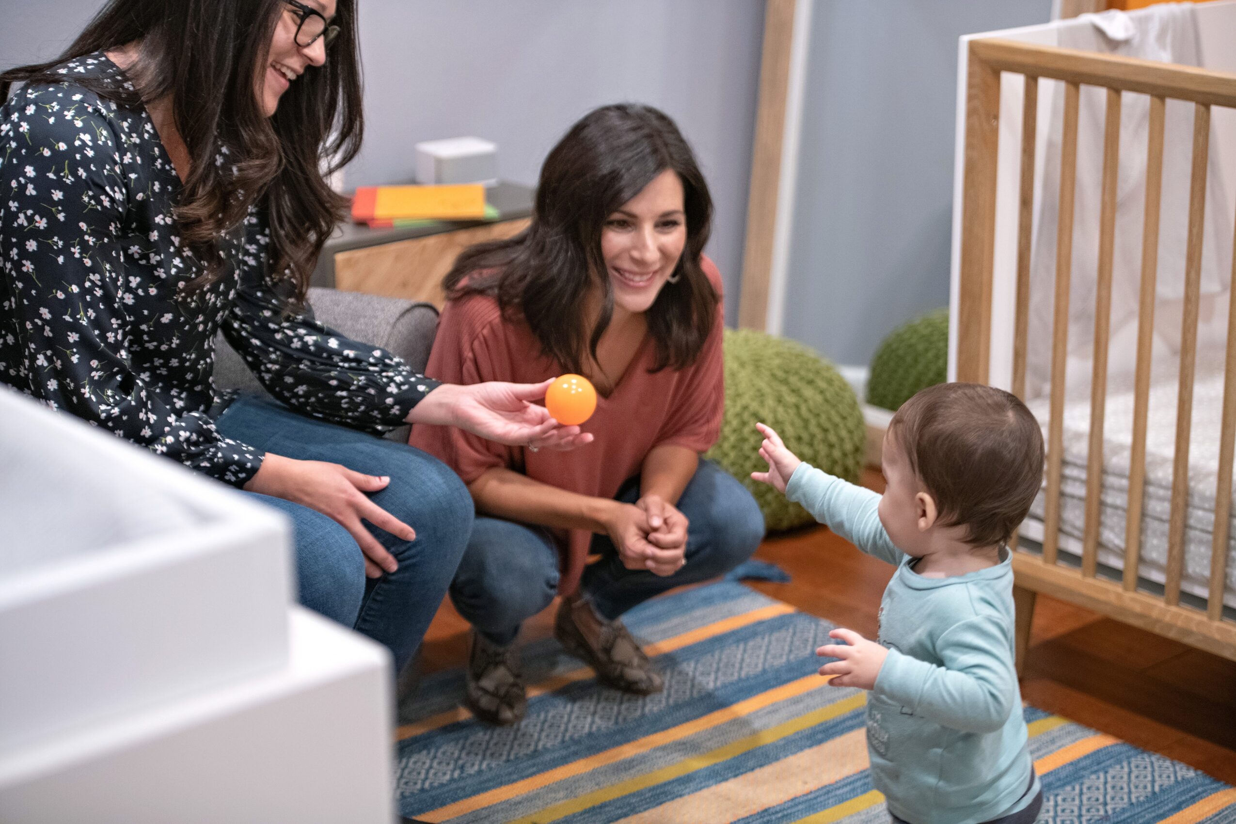 Two women and a young child playing with a ball indoors, near a crib and a colorful rug.