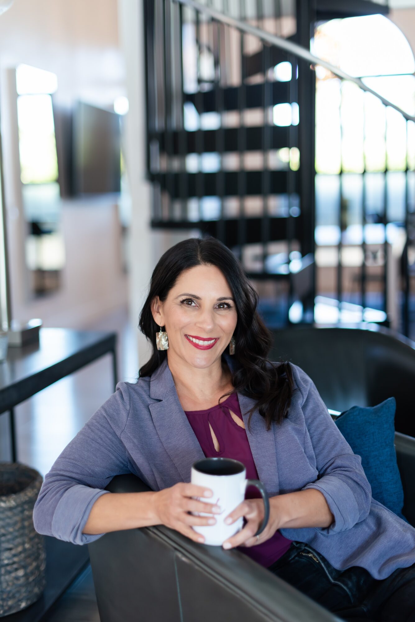 Woman with dark hair and earrings sitting on a sofa, holding a mug, smiling, in a modern interior.