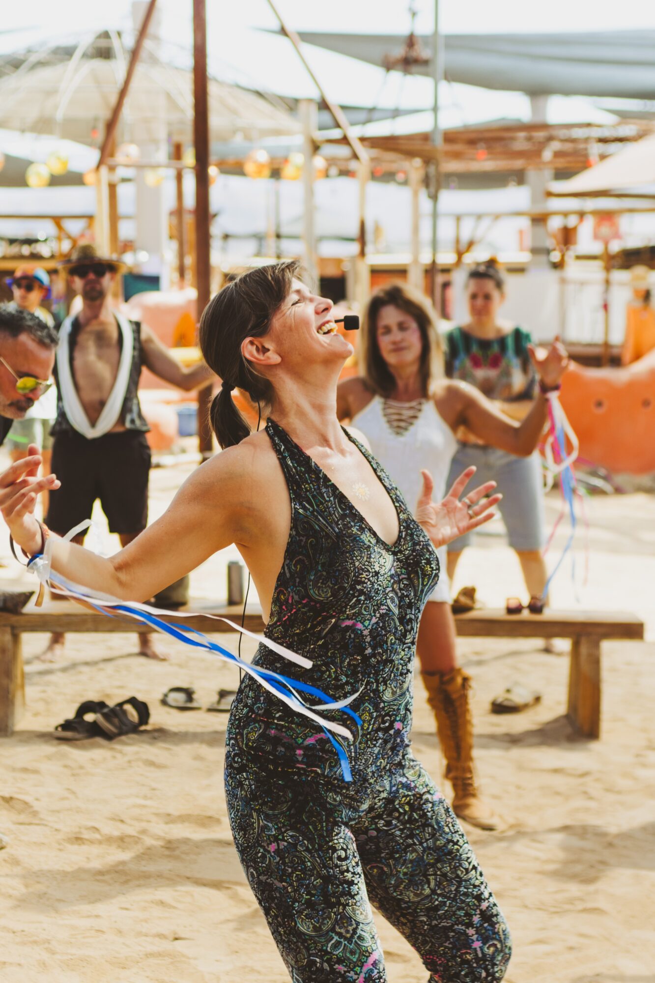 Woman in black jumpsuit smiling and dancing on sandy beach with people and boats in background.