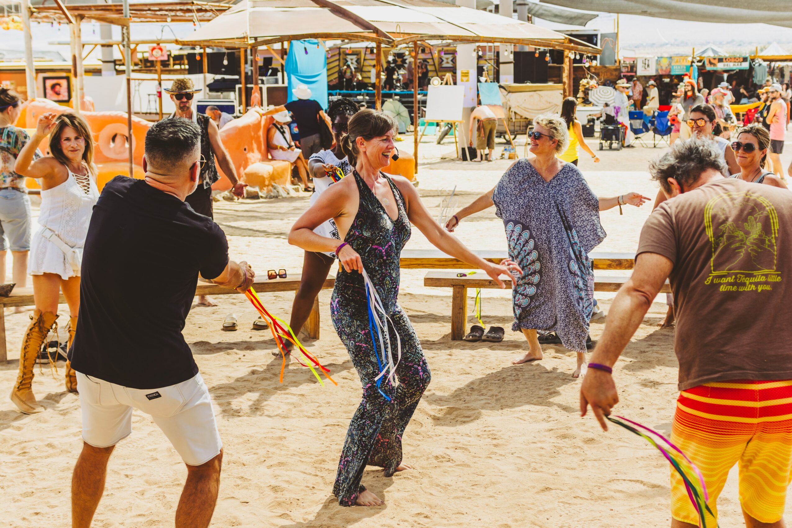 People dancing on sandy ground at an outdoor market or festival with stalls and tents in background.