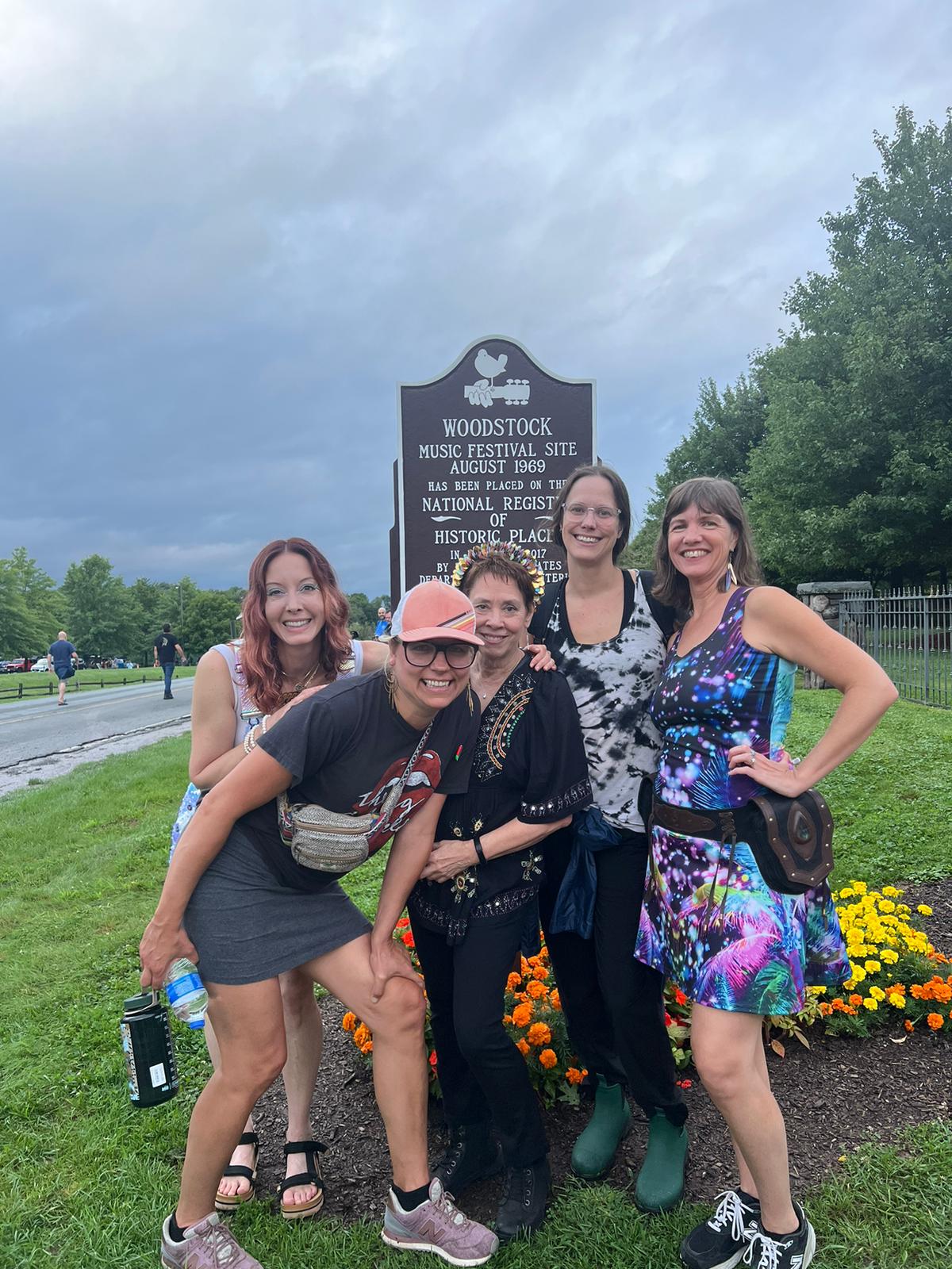 Five women standing outdoors near a historical marker, smiling, with trees and flowers in the background.