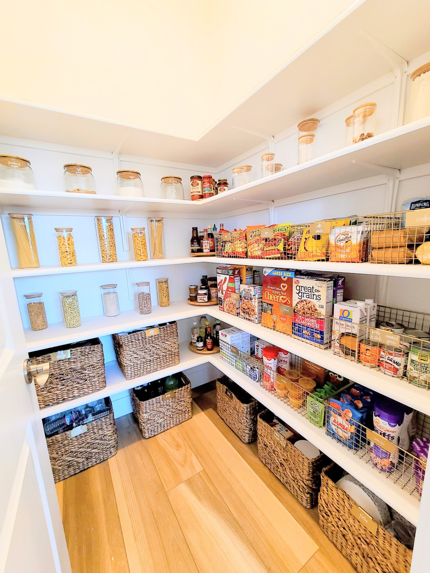 Pantry with shelves filled with jars, boxes, and baskets of food items, organized neatly, with a wooden floor.