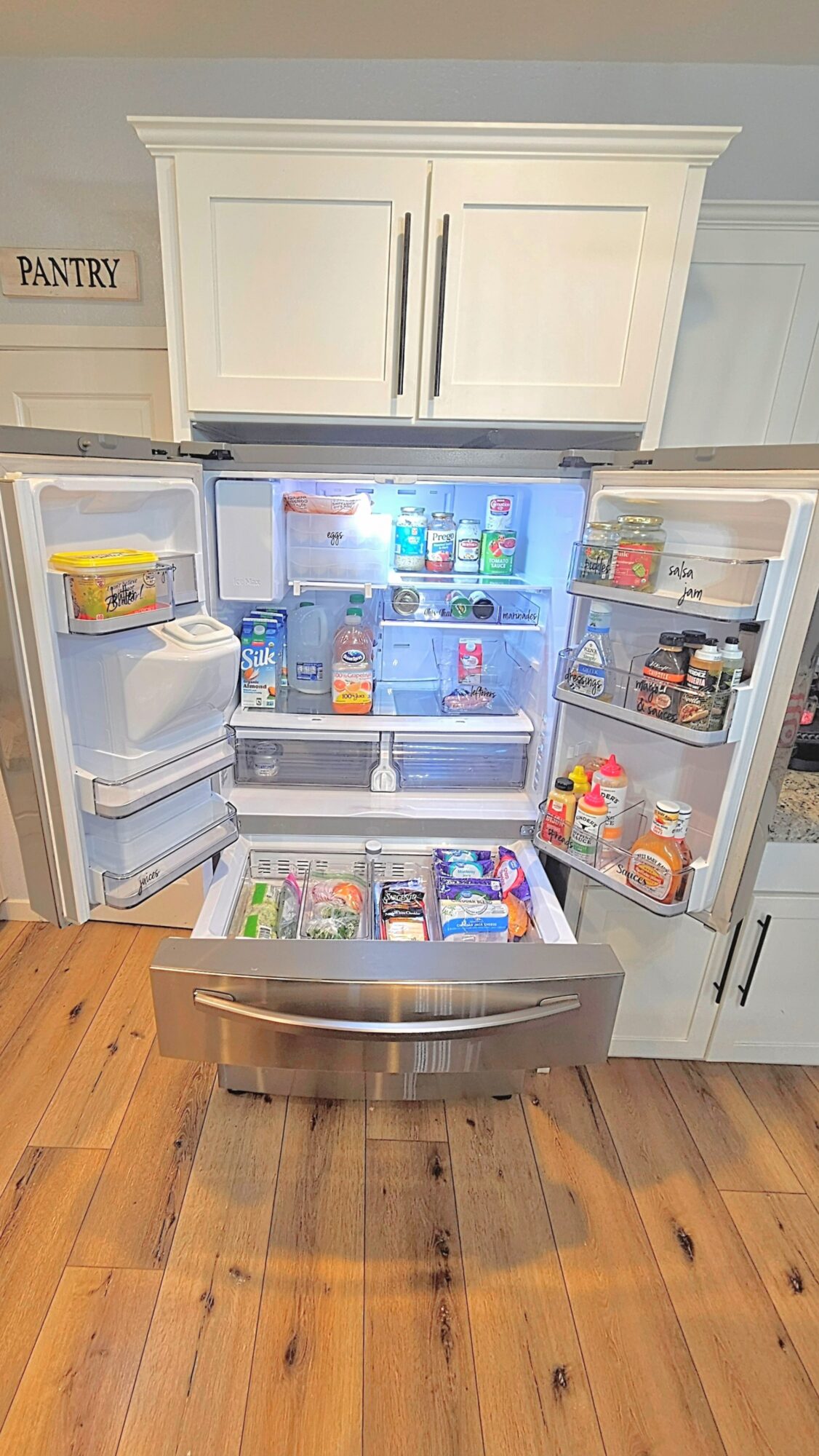 Open refrigerator with food items inside, kitchen cabinets above, and a pantry sign on the wall, wooden floor below.