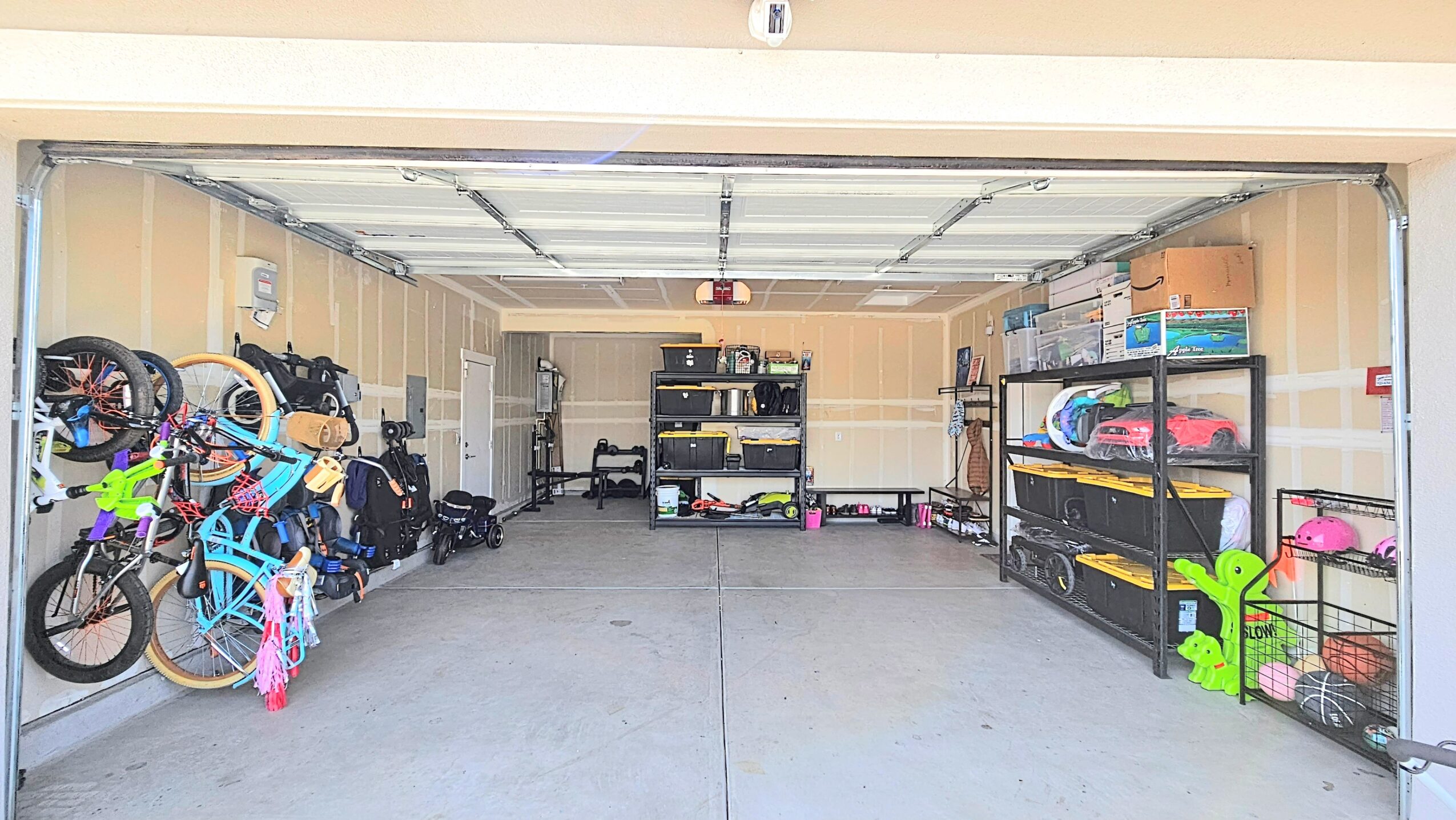 Garage with bicycles on the left, shelves with storage boxes and items on the right, and a workbench at the back.