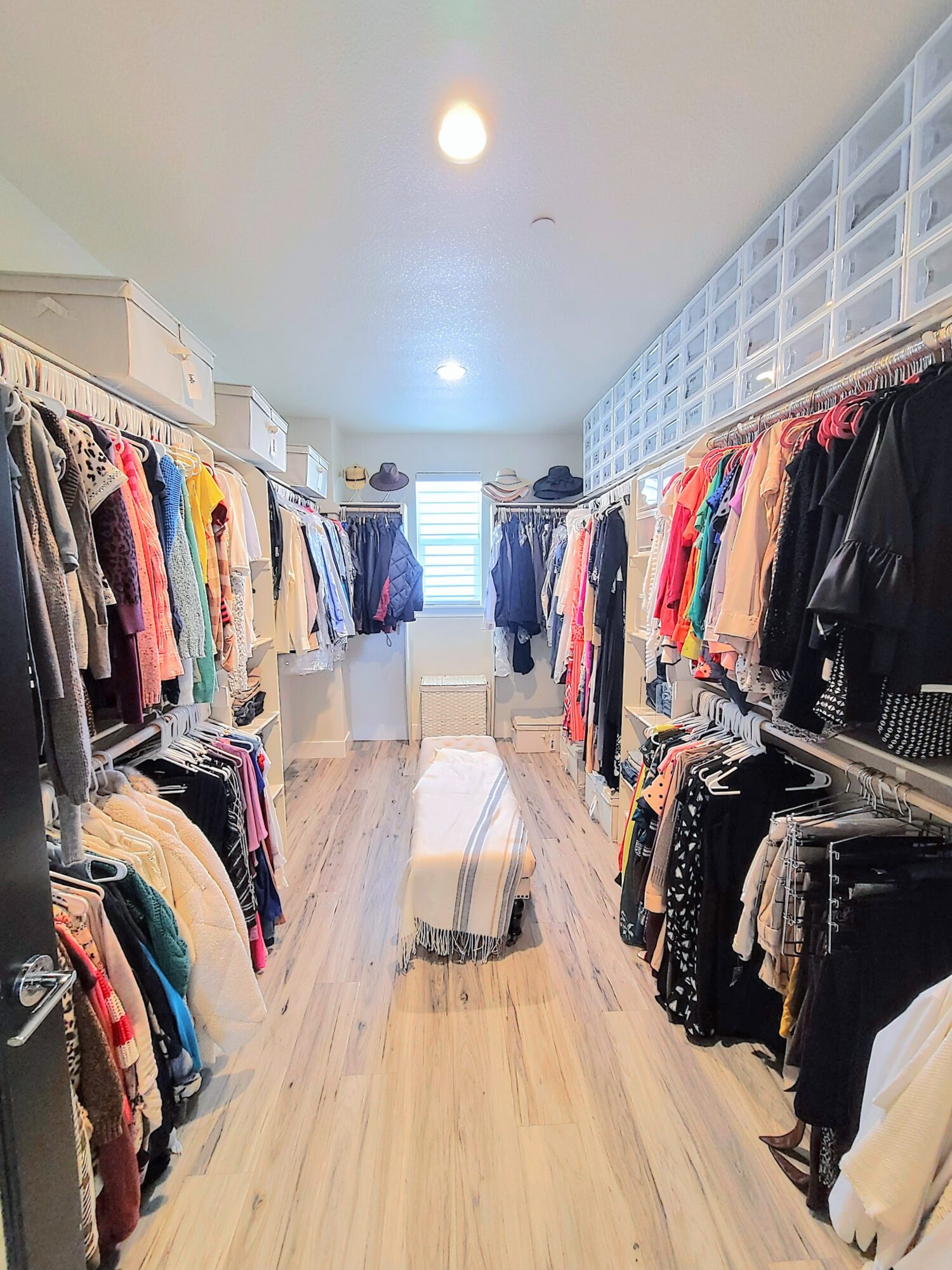 Clothing store aisle with racks of colorful clothes on both sides, a striped rug in the center, and a window at the end.
