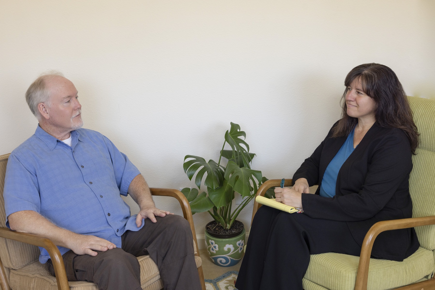 Older man and woman in a conversation, sitting in chairs with a potted plant between them, against a plain wall.