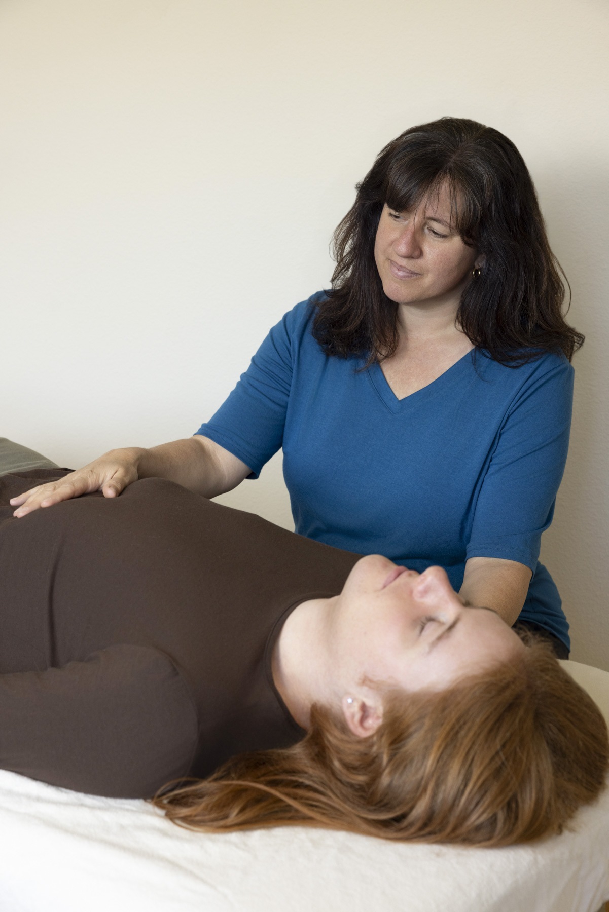 Woman in blue shirt performs a medical or massage procedure on a lying woman with red hair, in a clinical setting.