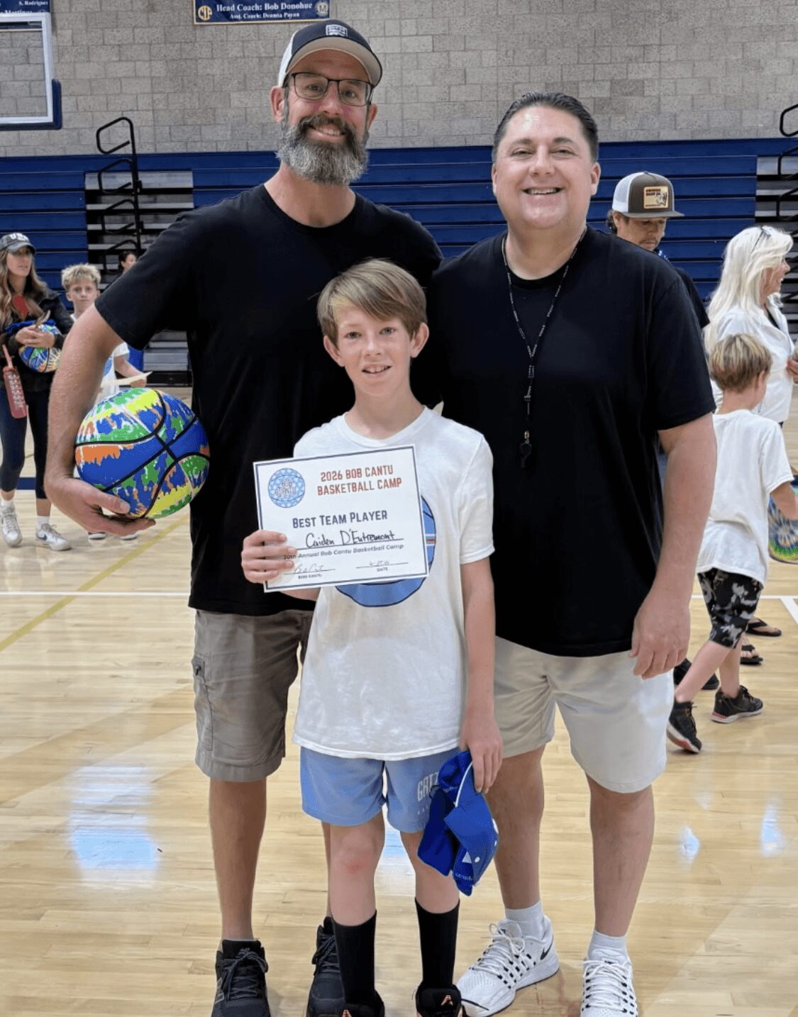 Three people standing in a gymnasium, a young boy holding a certificate, with two adults smiling beside him.