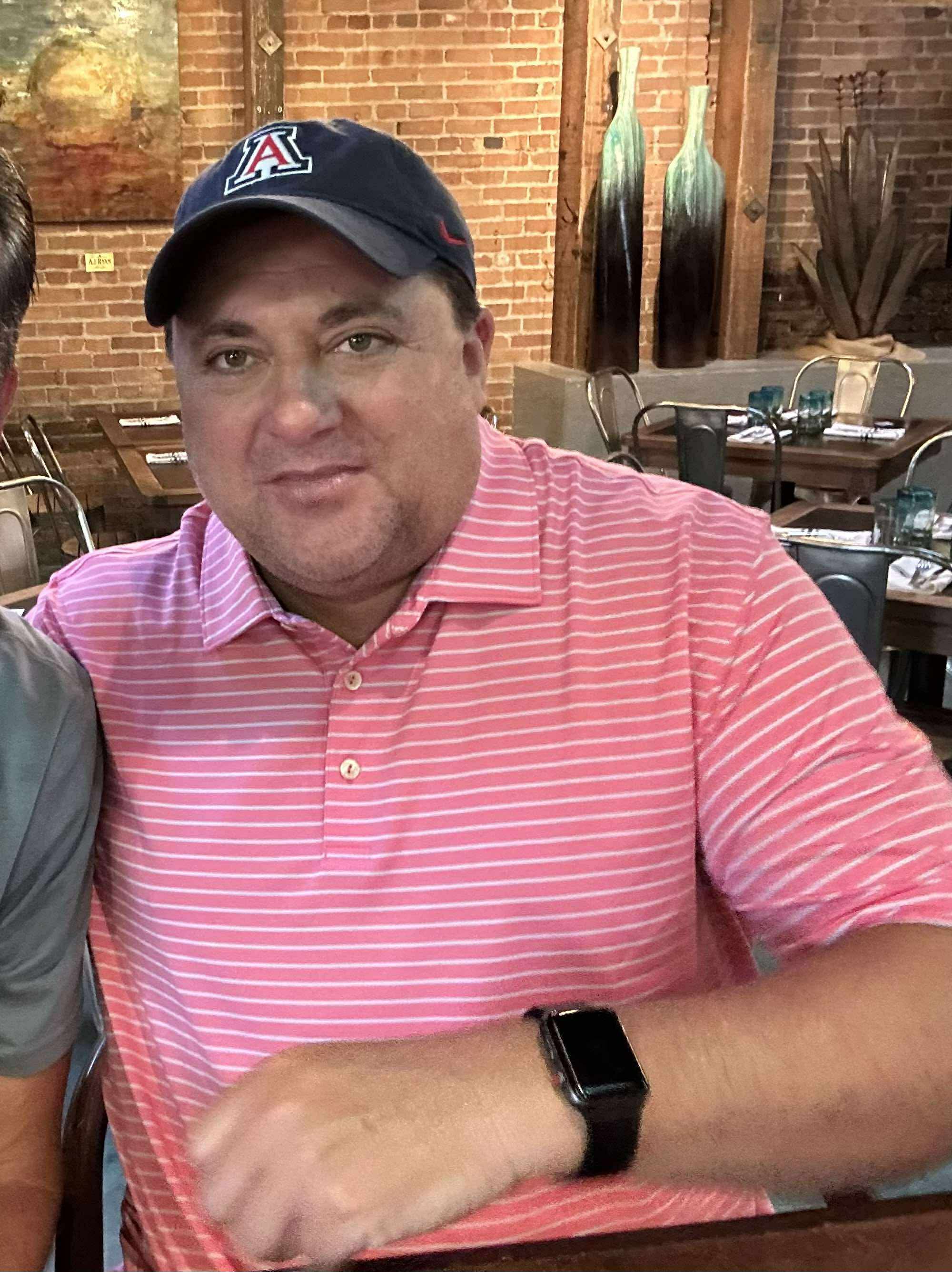 Man wearing a pink striped polo shirt, black cap, and smartwatch, sitting at a table in a restaurant with brick walls and decor.